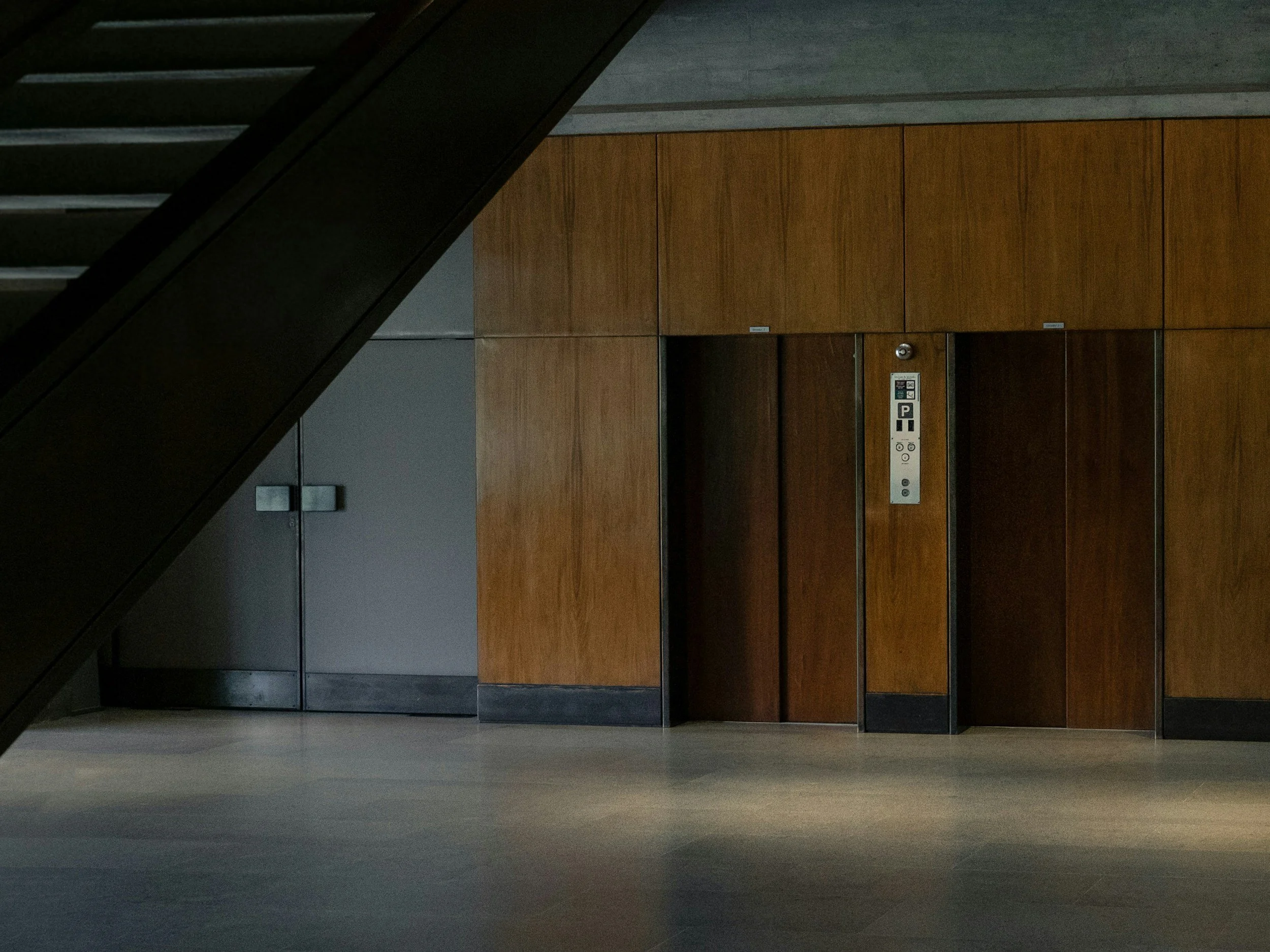 Photo of an elevator door with dark wood paneling and a control panel, situated under a staircase in a modern building.