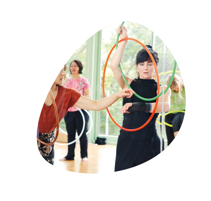 HulaHoop Dance Workshop für Erwachsene, eine Frau hilft einer anderen Frau mit zwei bunten HulaHoop Reifen