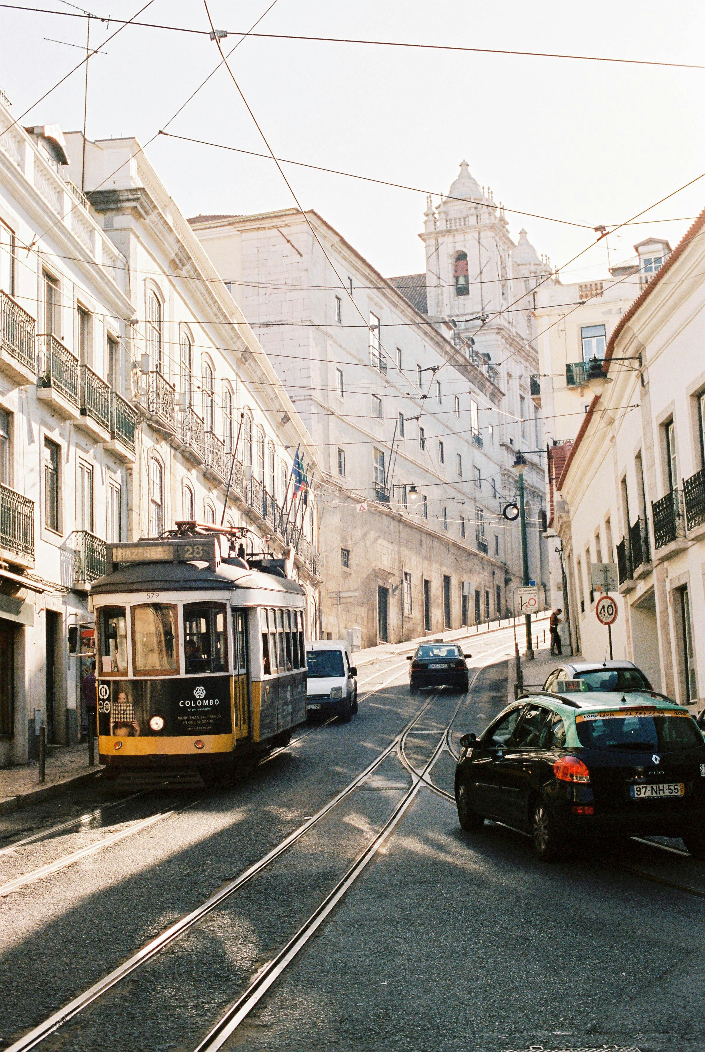 Imagem de uma rua com prédio antigo, com um bonde amarelo na frente e carruagens de carros estacionados, fios de energia cruzando o céu, e uma igreja ao fundo.