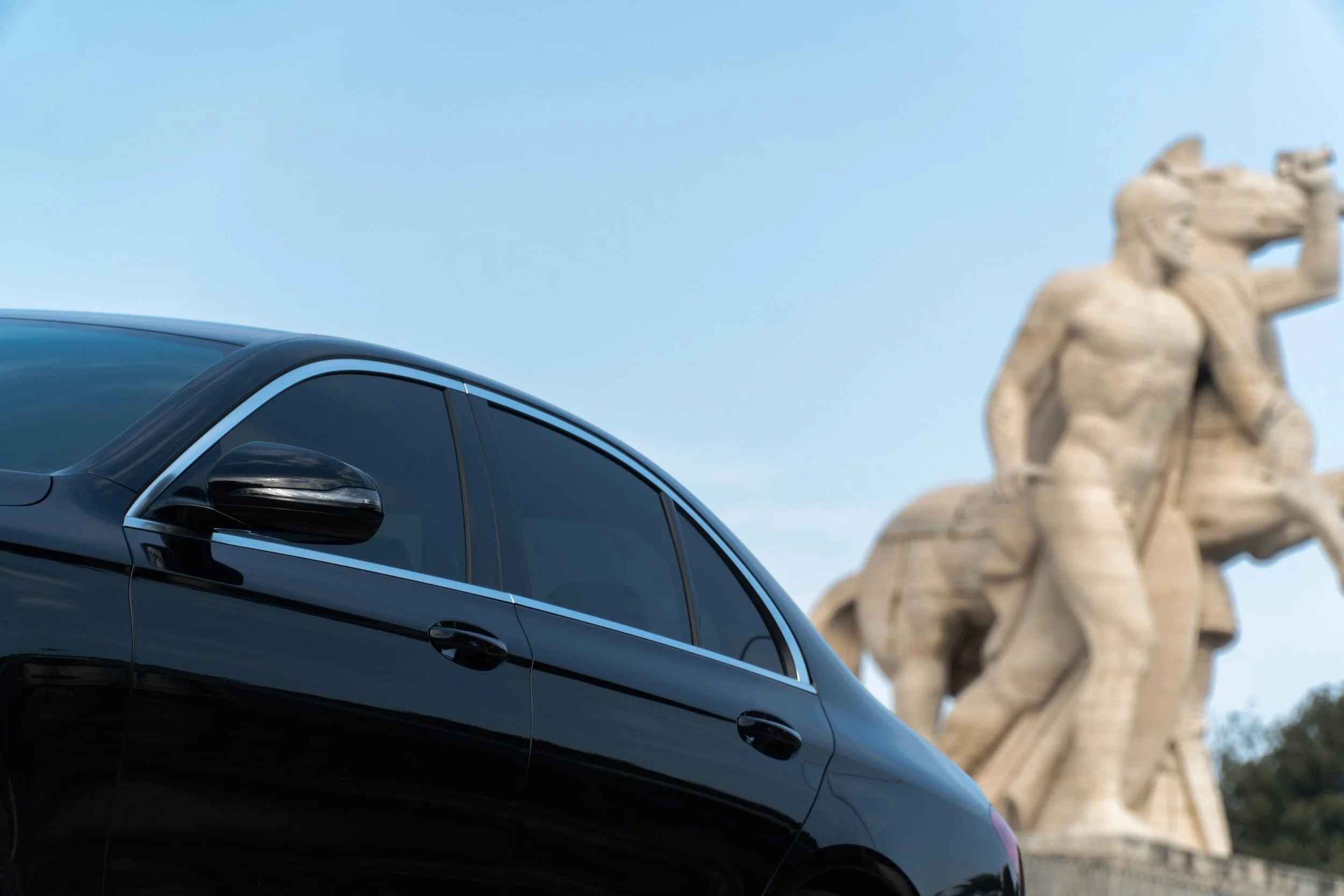 Carro preto reflete o céu azul claro, com uma estátua de dois alces em destaque ao fundo.