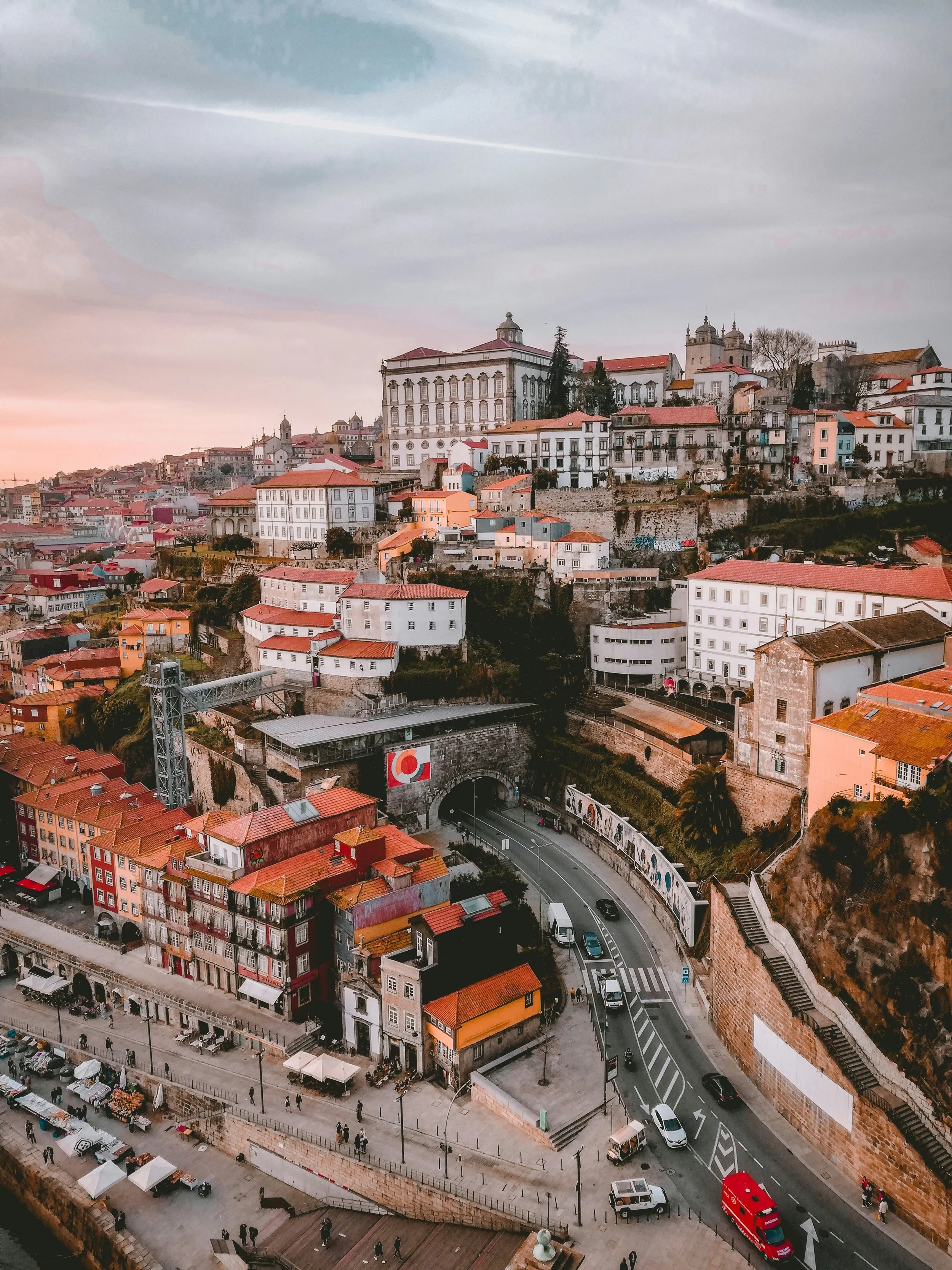 Vista de uma cidade com edifícios históricos, ruas movimentadas, transporte e muitos prédios com telhados vermelhos, em uma cena urbana ao entardecer.
