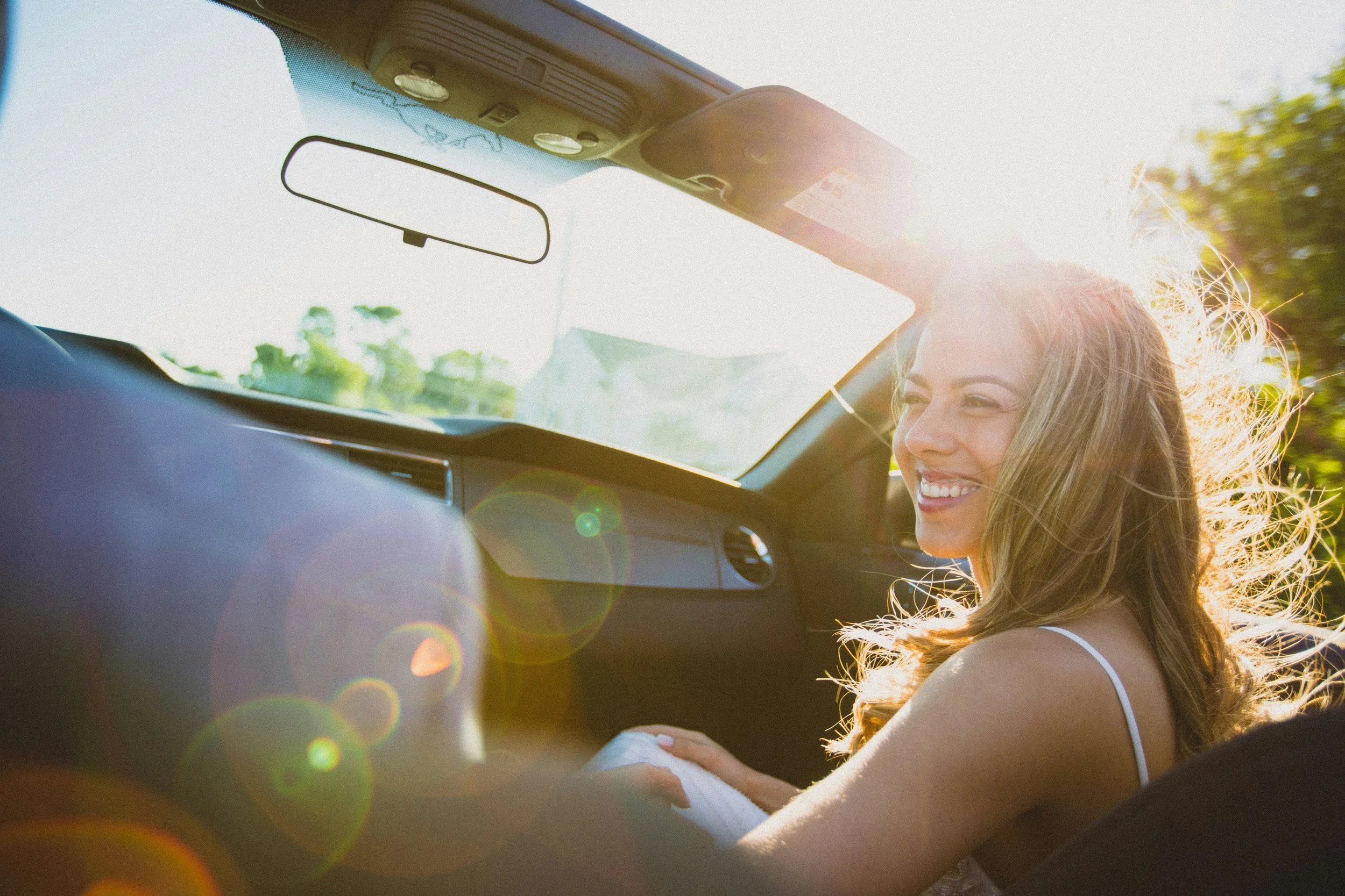 Mulher sorridente sentada no banco do passageiro de um carro, com luz do sol brilhando e árvores ao fundo.
