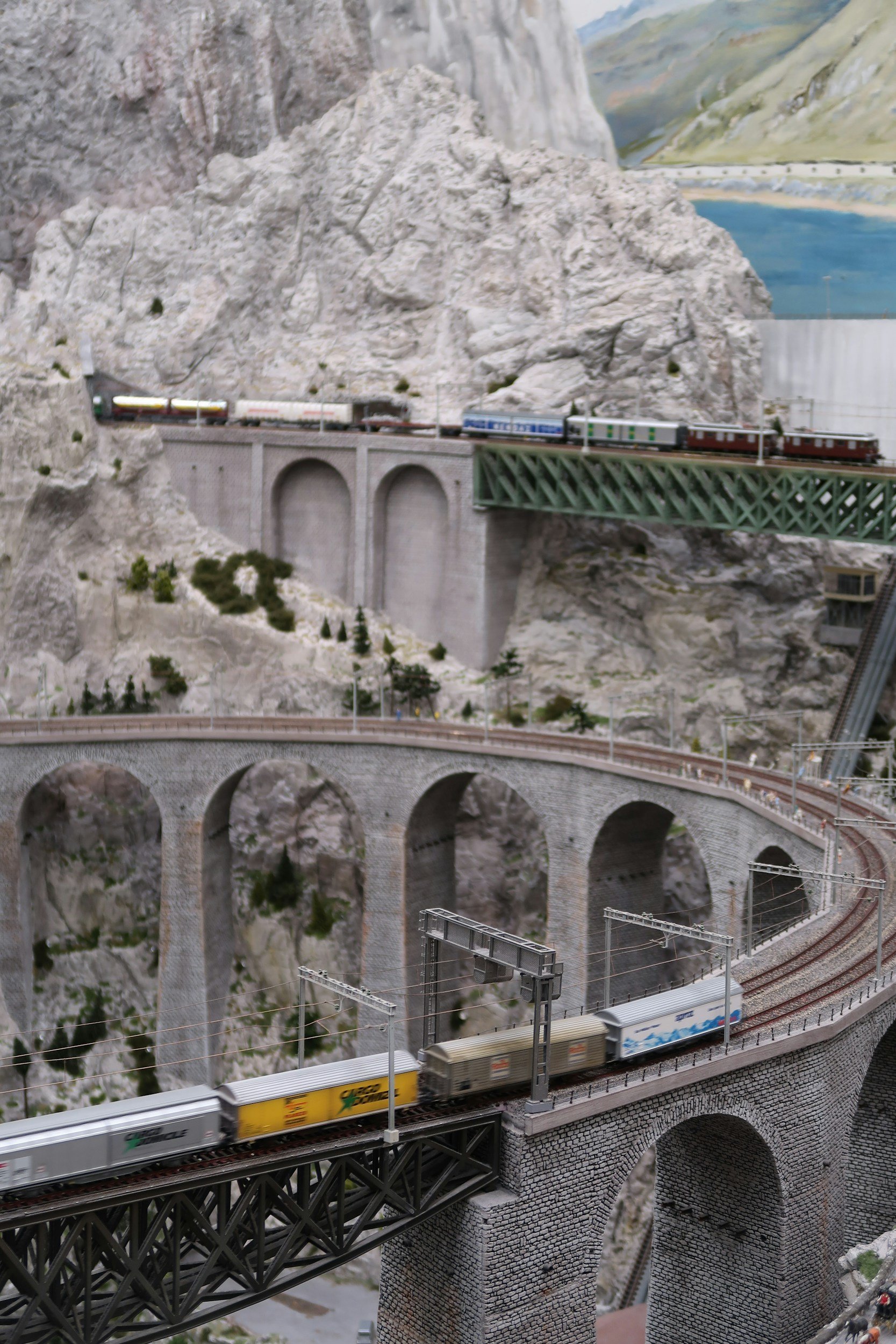 A detailed model railway scene set in a mountainous landscape with a train crossing a stone arched bridge, with mountains, valley, and a painted backdrop of mountains and water in the distance.