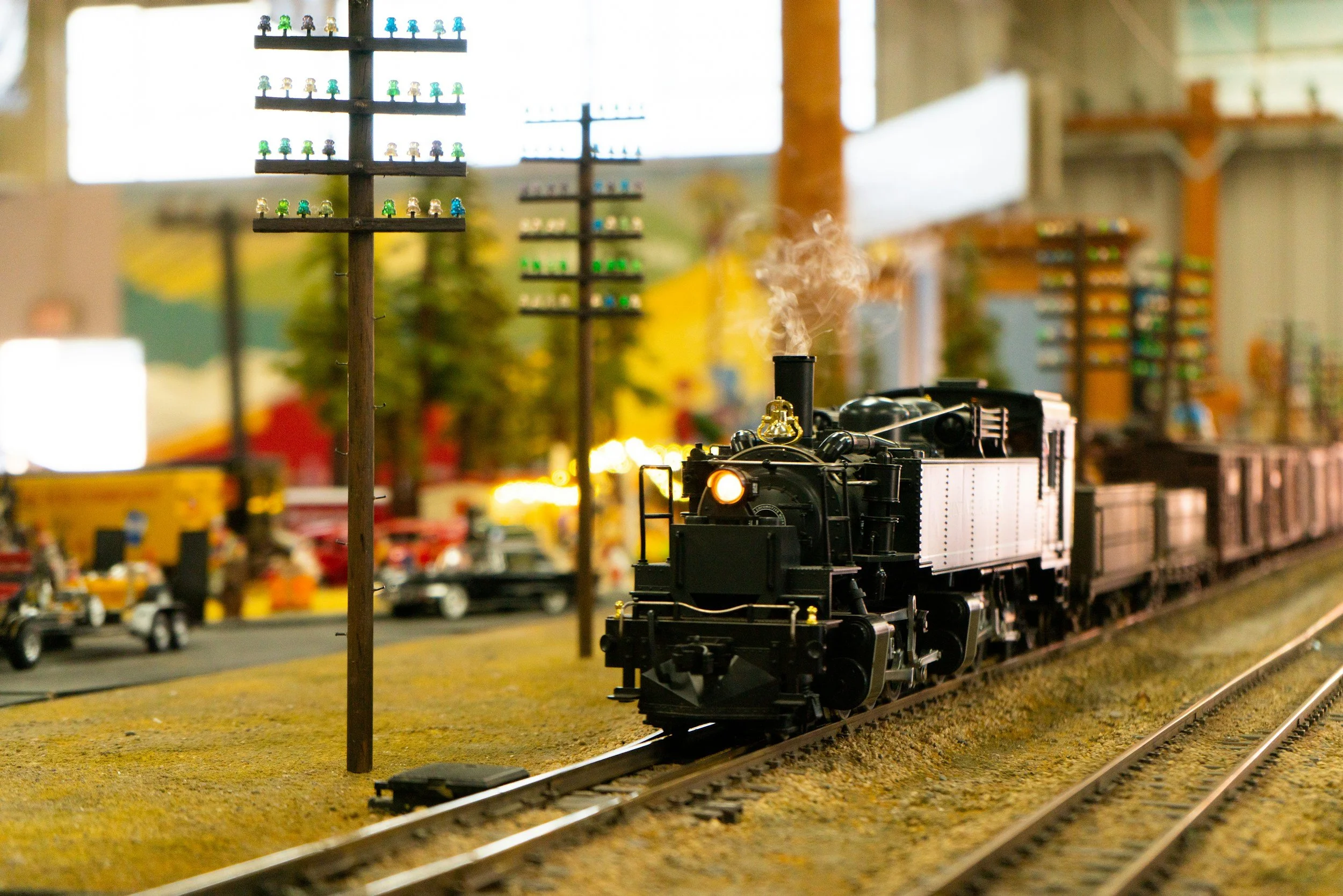 Model train with black steam locomotive on tracks, small signal pole, and colorful trees in the background.
