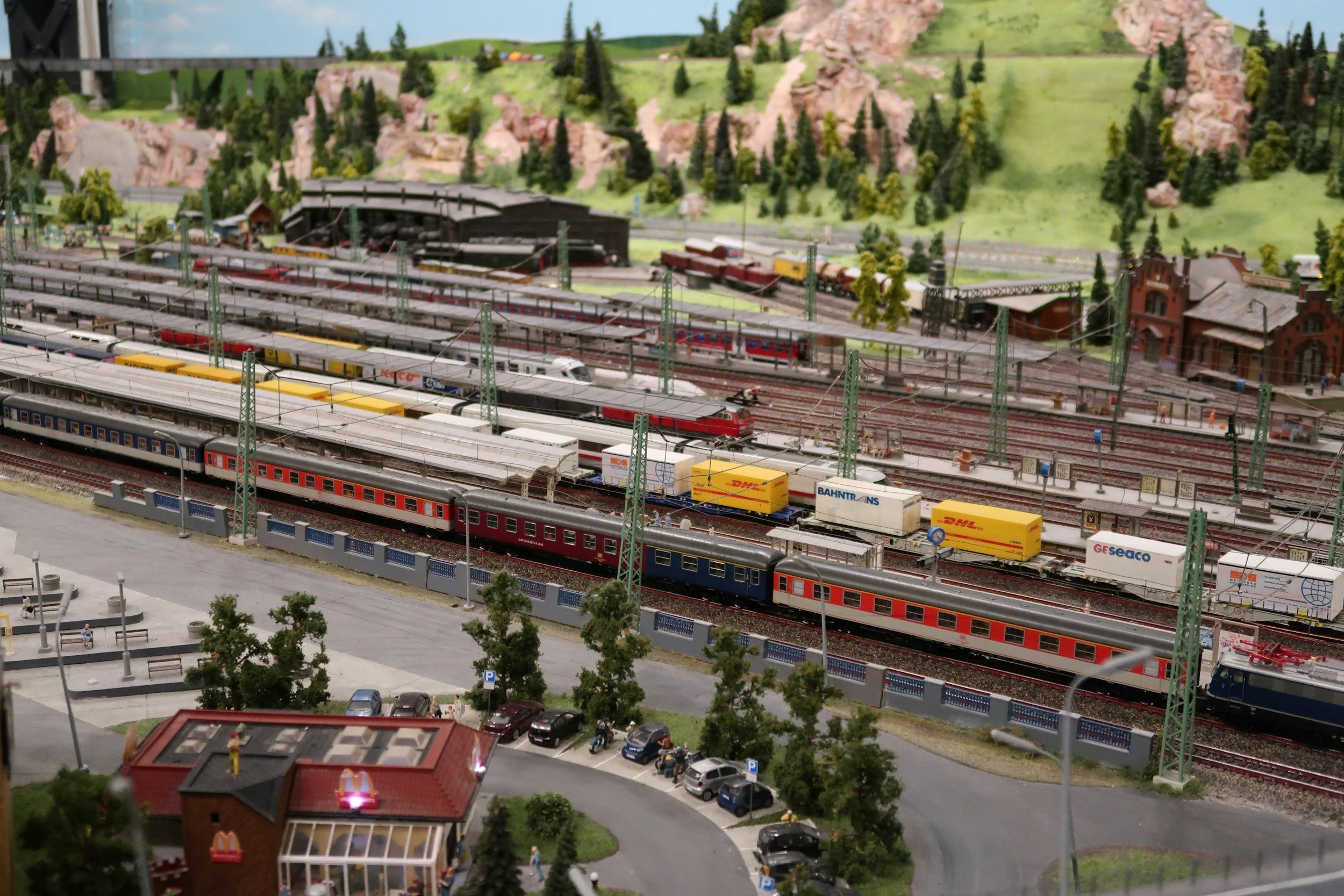 A detailed model train layout featuring multiple train tracks with various model trains, a scenic backdrop with mountains and trees, a small station, and surrounding miniature buildings and vehicles.