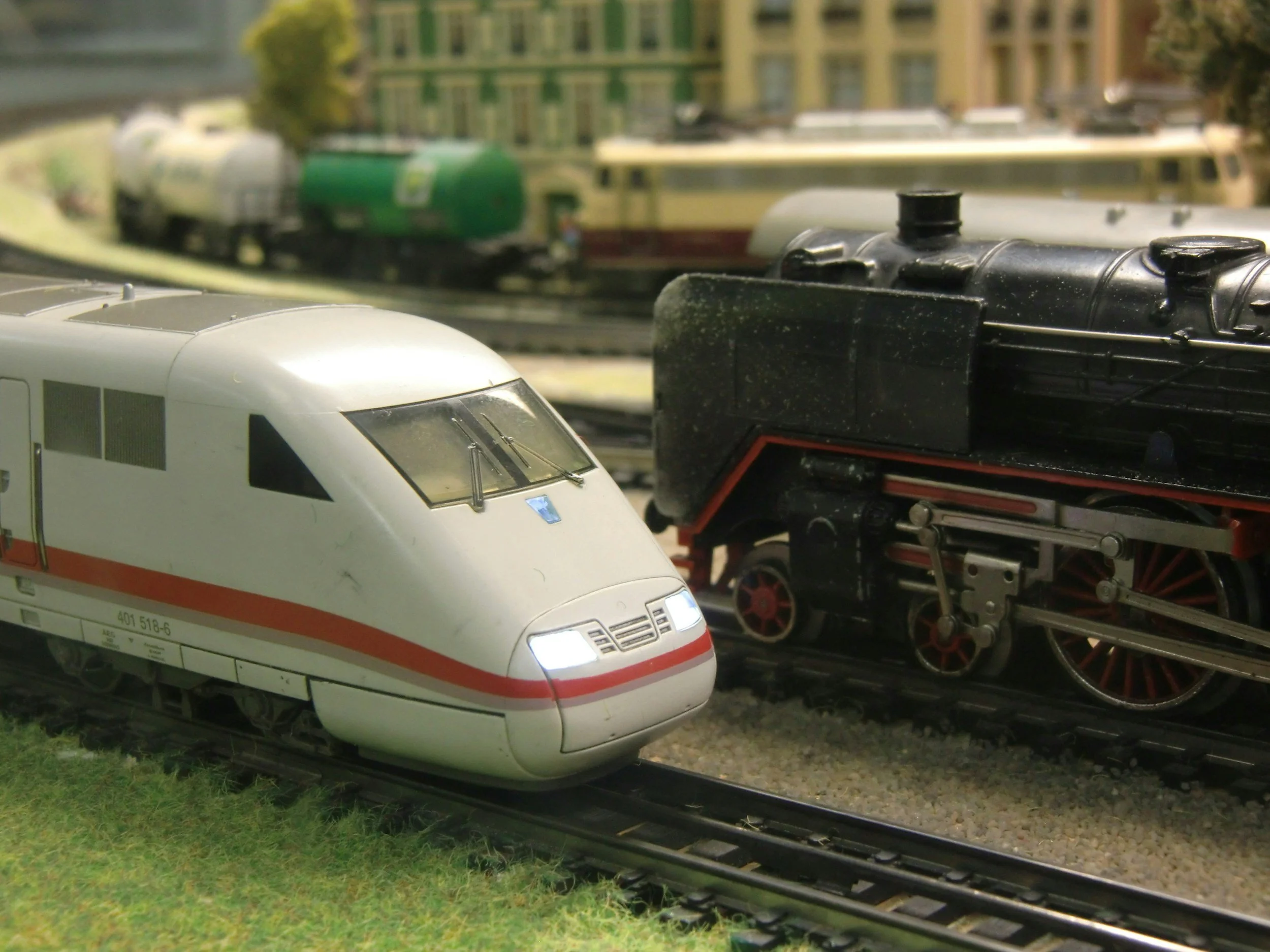 Model train set with a white high-speed train in the foreground and a black steam engine in the background on a miniature railroad track with buildings and trees in the distance.