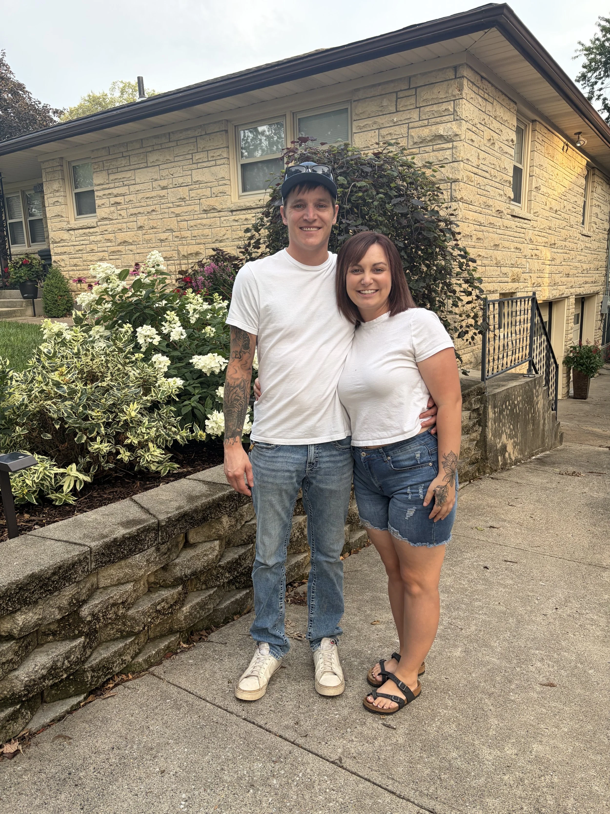 A smiling man and woman standing close together outdoors in front of a beige brick house with garden bushes and flowers. The man is wearing a white t-shirt, jeans, and a baseball cap, while the woman is dressed in a white t-shirt and denim shorts.