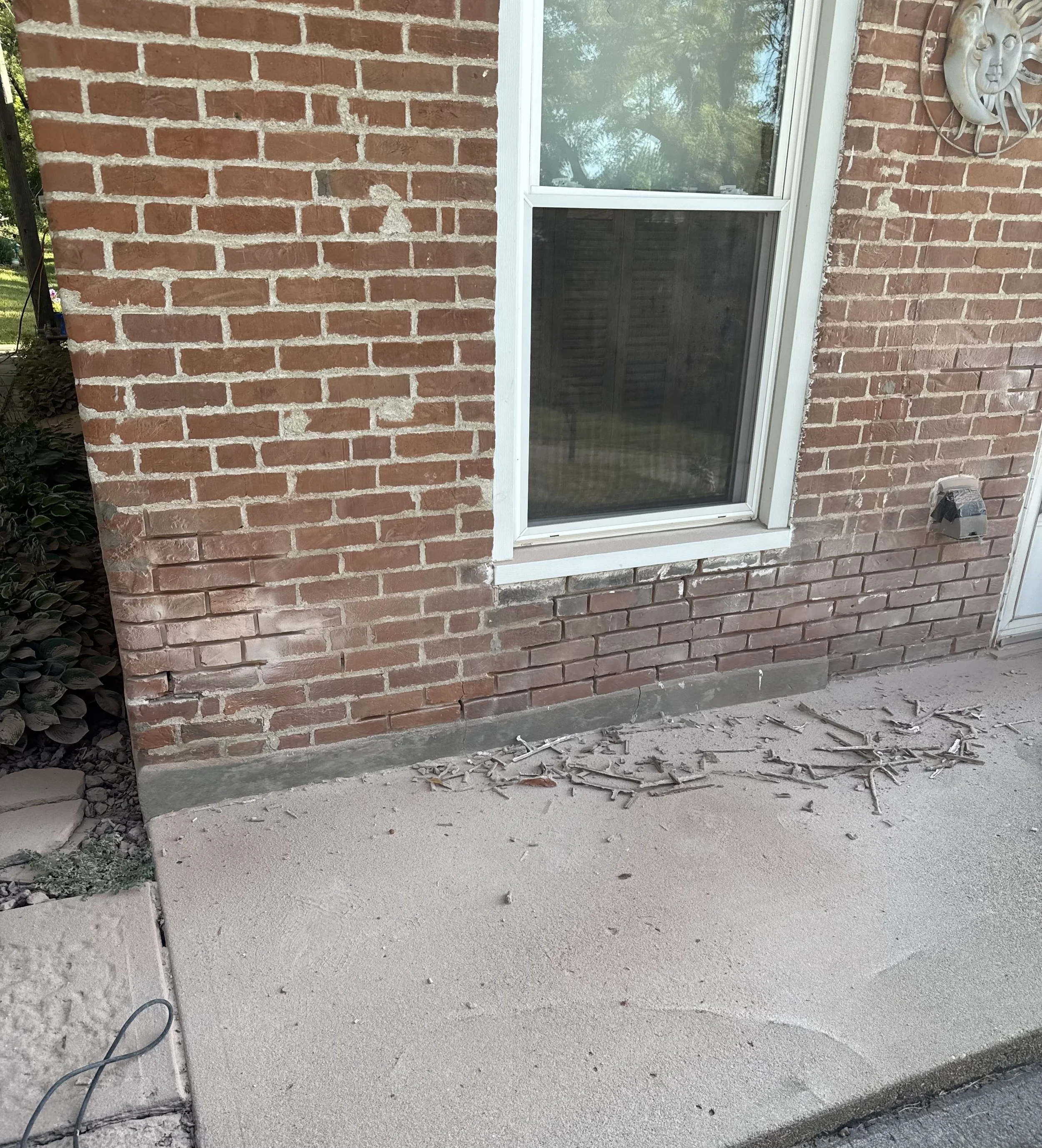 Close-up of a brick building wall with a multi-pane window and decorative sun face wall art. Part of a sidewalk with concrete and gravel is visible in the foreground.