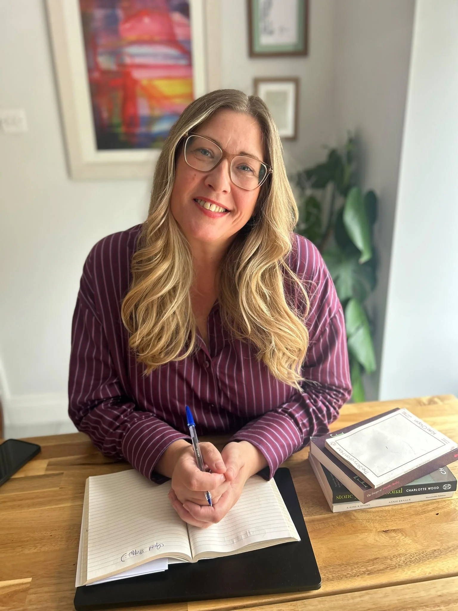 A woman with long blonde hair, wearing glasses and a striped maroon shirt, smiling while sitting at a wooden table in a room with framed artwork on the wall and a potted plant in the background. She is writing in an open notebook with a blue pen, surrounded by a stack of books and a laptop.