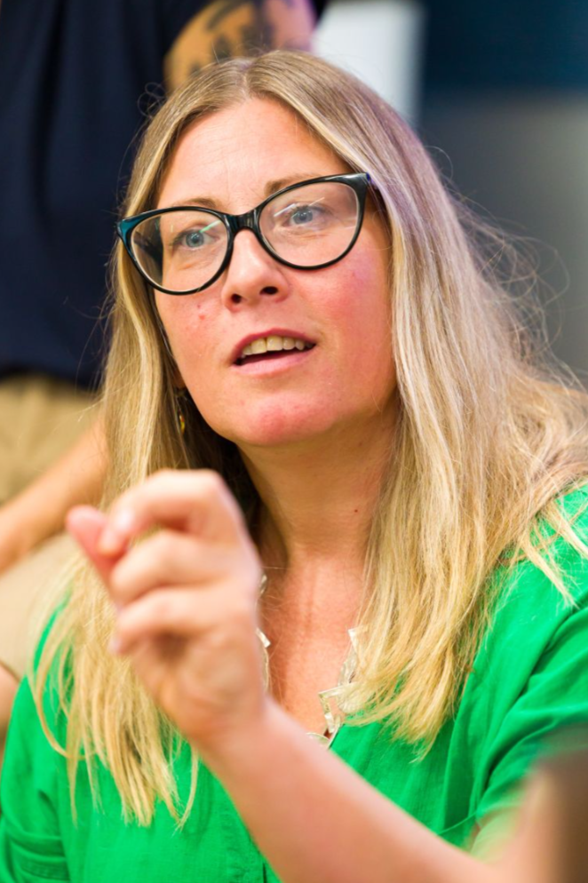 A woman with long blonde hair, glasses, and a green shirt, gestures with her hand while speaking.