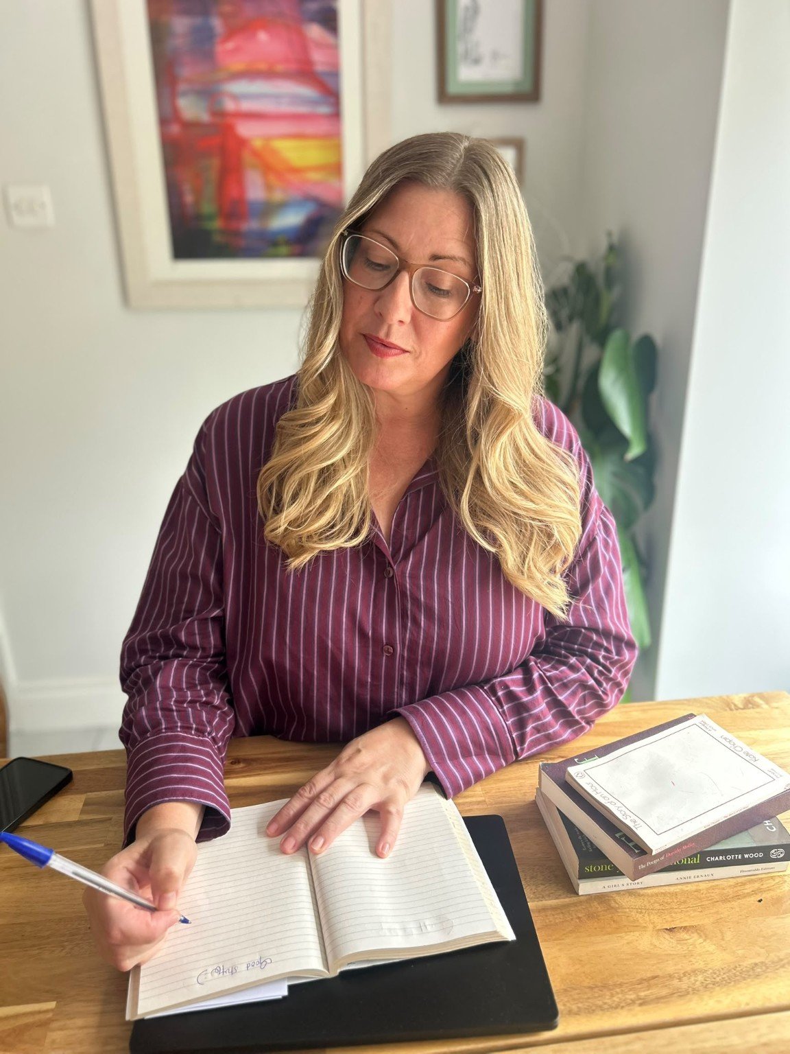 Woman with blonde hair and glasses writing in a notebook at a wooden desk, with a stack of books nearby.