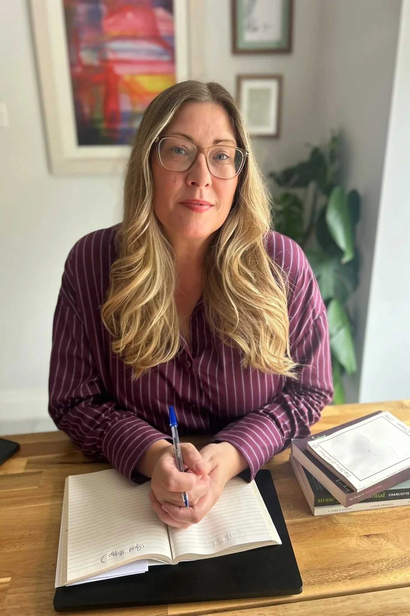 A woman with long blonde hair wearing glasses and a striped maroon shirt sitting at a wooden table, writing in a notebook with a blue pen. There are books and papers on the table, and a plant and framed pictures in the background.