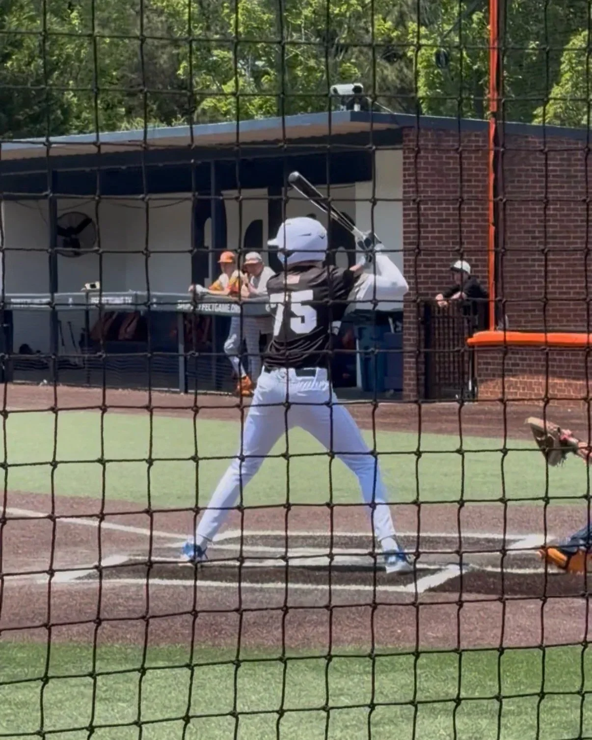 A baseball player in uniform is at bat, preparing to swing at a pitch on a baseball field. The scene is viewed through a black protective net, with a barbecue area and two people in the background.