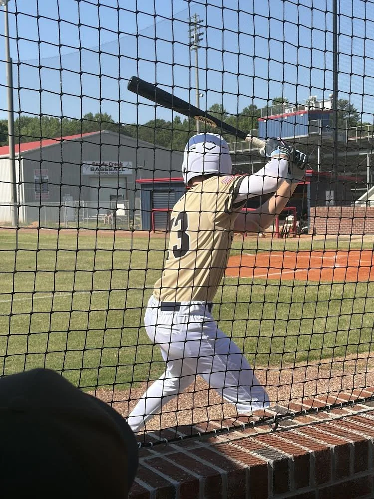 A baseball player in a beige jersey and white pants practicing batting inside a batting cage, with the fence and a baseball field in the background.