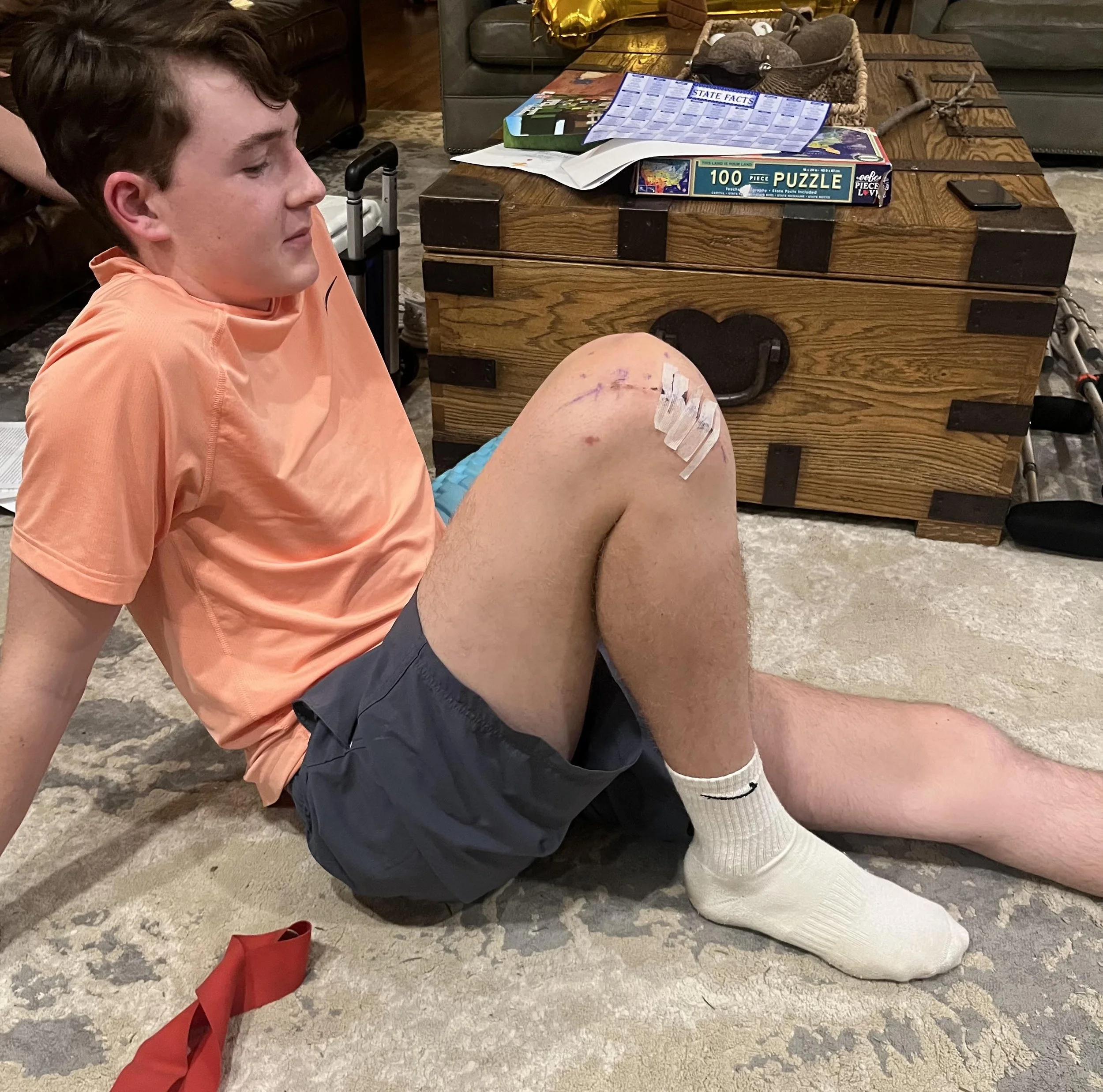 A young man with brown hair is sitting on the floor with his knee bandaged and medical tape on his thigh. He is wearing an orange T-shirt, black shorts, and white knee-high socks. The scene likely depicts a medical or injury situation.
