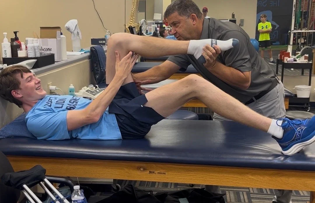 A physical therapist helping a young man with a leg exercise in a medical or sports clinic, with gym equipment and other people in the background.