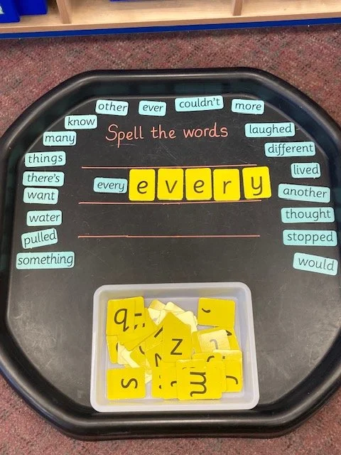 A classroom activity with a black tray, a container of yellow letter tiles, and a card that says 'Spell the words'. The activity involves forming the word 'every' using letter tiles.