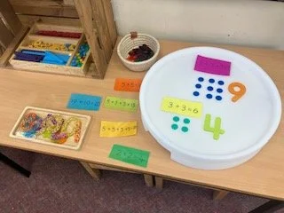 Educational math activity setup with a large white circle labeled with the number 9, colorful flashcards with math problems and answers, and small bowls containing counters and beads for counting or solving math problems.