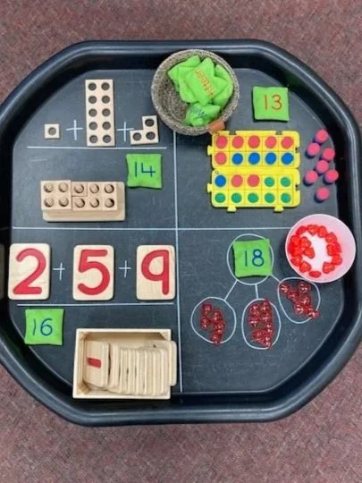 Educational math and counting activities for children on a black tray. Includes dominoes, a basket of green socks, a yellow and red dot game, pink beads, a green block with the number 13, a purple block with the number 14, red beads with the number 18, and a wooden abacus.