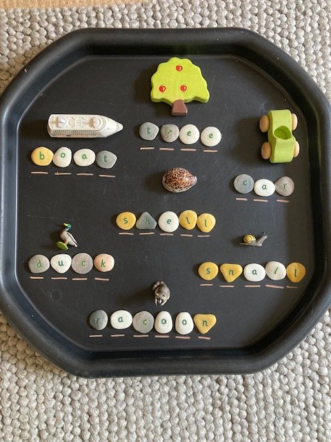 A black tray with foam letter beads spelling words like boat, tree, car, duck, snail, and ocean. The tray also has small toys and objects, including a toy boat, a tree shaped eraser, a small rock, a tiny snail, and a small penguin figure.