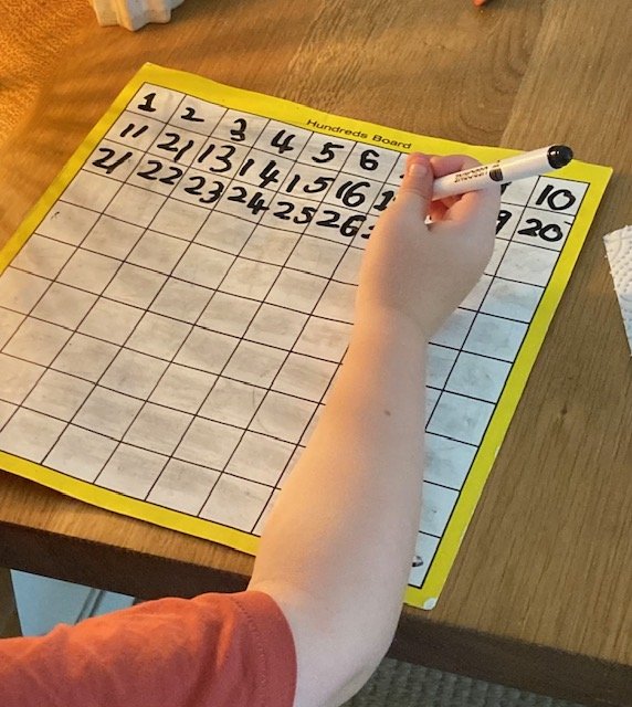 A person writes on a yellow hundred board with numbered squares from 1 to 100, filling in the first three rows with numbers, on a wooden surface.