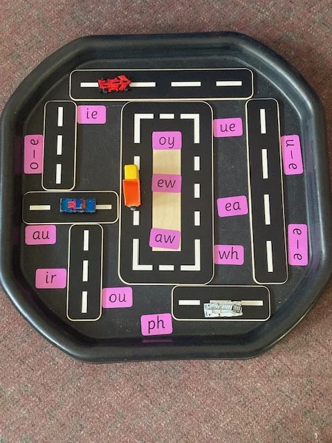 A tabletop game resembling a mini road map with various colored magnetic or plastic vehicles and pink letter tiles with letter combinations.