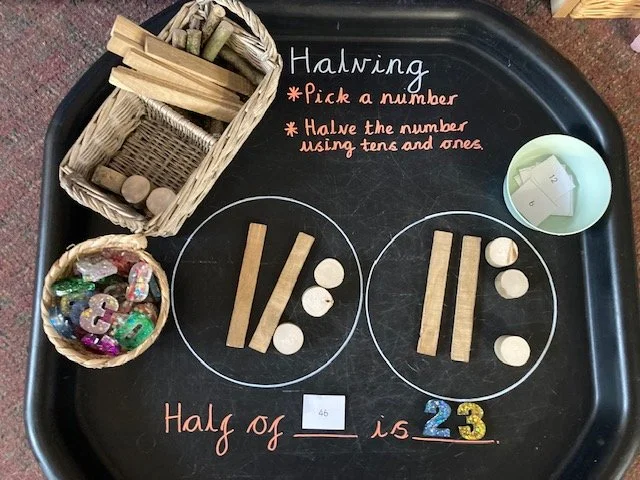 A black tray with educational math tools for teaching halving, including wooden sticks, counters, base ten blocks, and paper number cubes. Instructions for halving by using tens and ones are written on the tray.