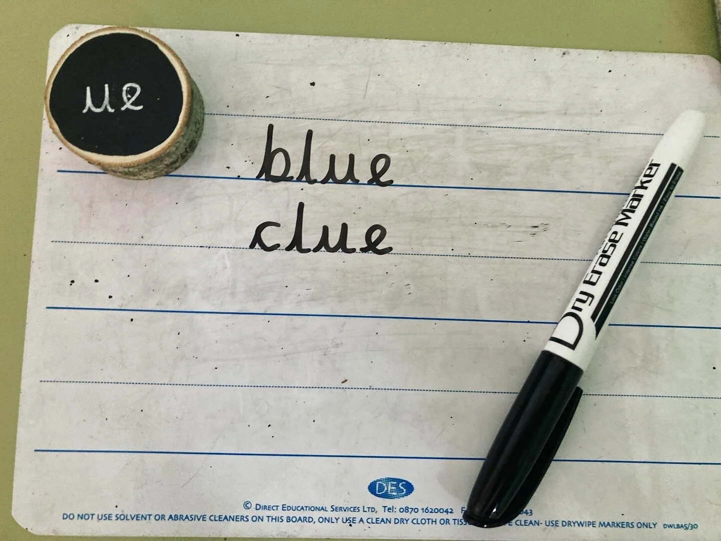 A whiteboard with the word "blue" written twice in English with black markers, a black marker lying on the board, and a small round chalkboard with the word "Blue" written in cursive, placed at the top left corner.