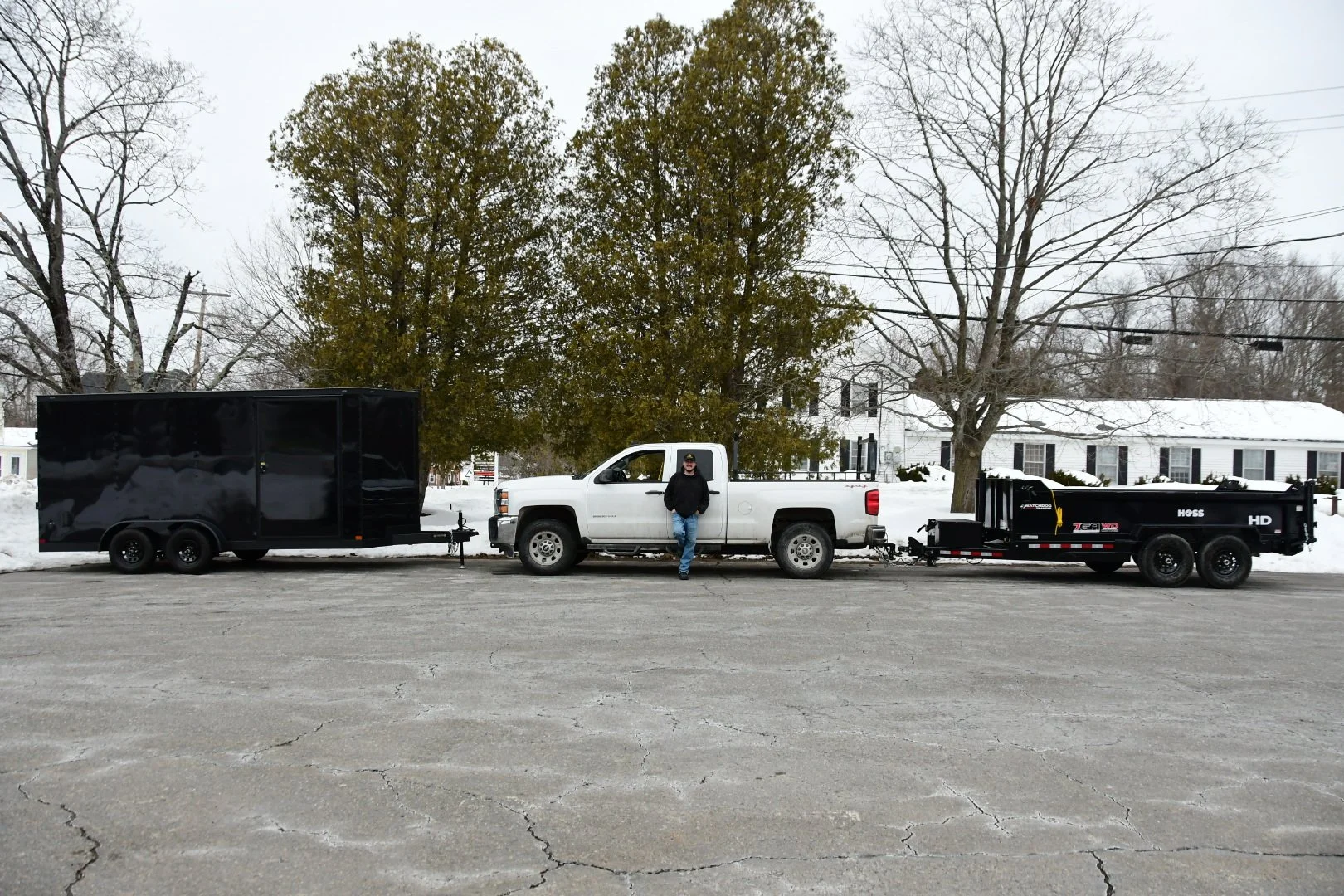 A man standing next to a white pickup truck attached to a black trailer and a black flatbed trailer on a snowy street with trees and houses in the background.