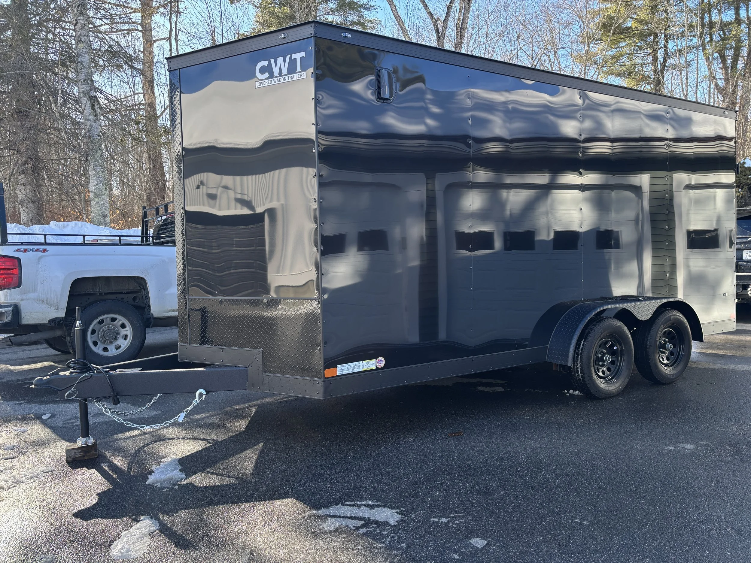 A black enclosed trailer attached to a white pickup truck parked on an asphalt surface with remnants of snow. The trailer has the logo "CWT" for Covered Wagon Trailers and is surrounded by leafless trees and a bright sky.