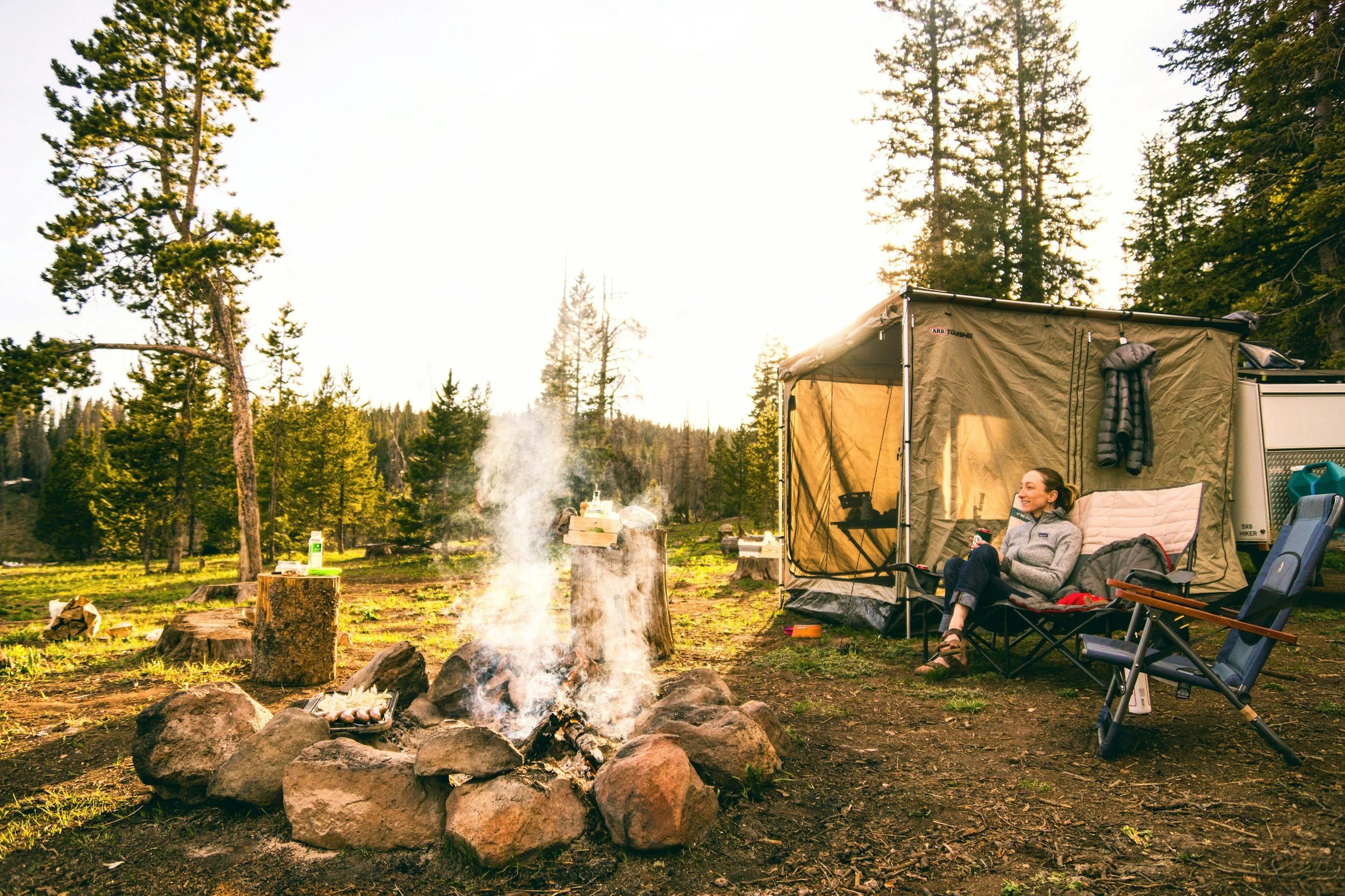 A woman sitting and relaxing in camping chairs near a campfire in a forested area with tall pine trees during sunset or sunrise.
