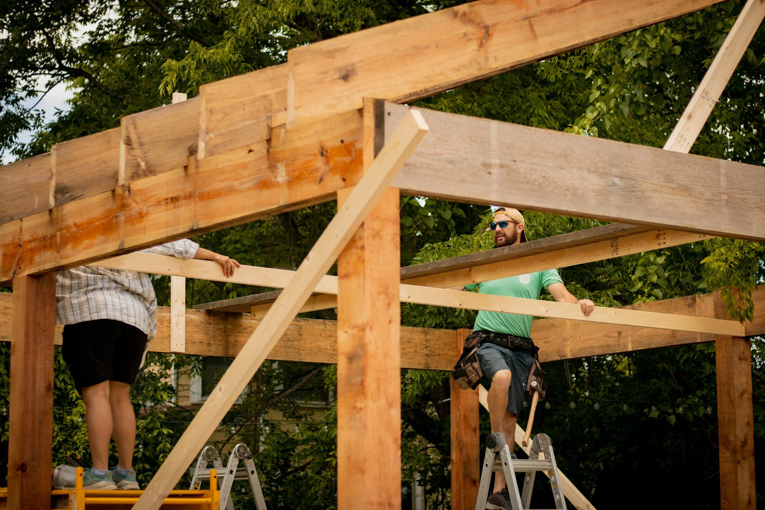 Two men are building a wooden deck outdoors, supported by ladders and surrounded by green trees.