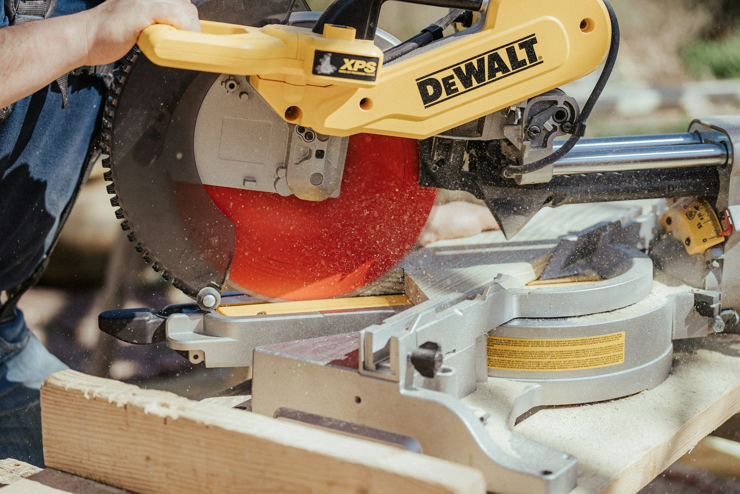 A person operating a yellow DEWALT circular saw on a piece of wood outdoors, with sawdust flying around.
