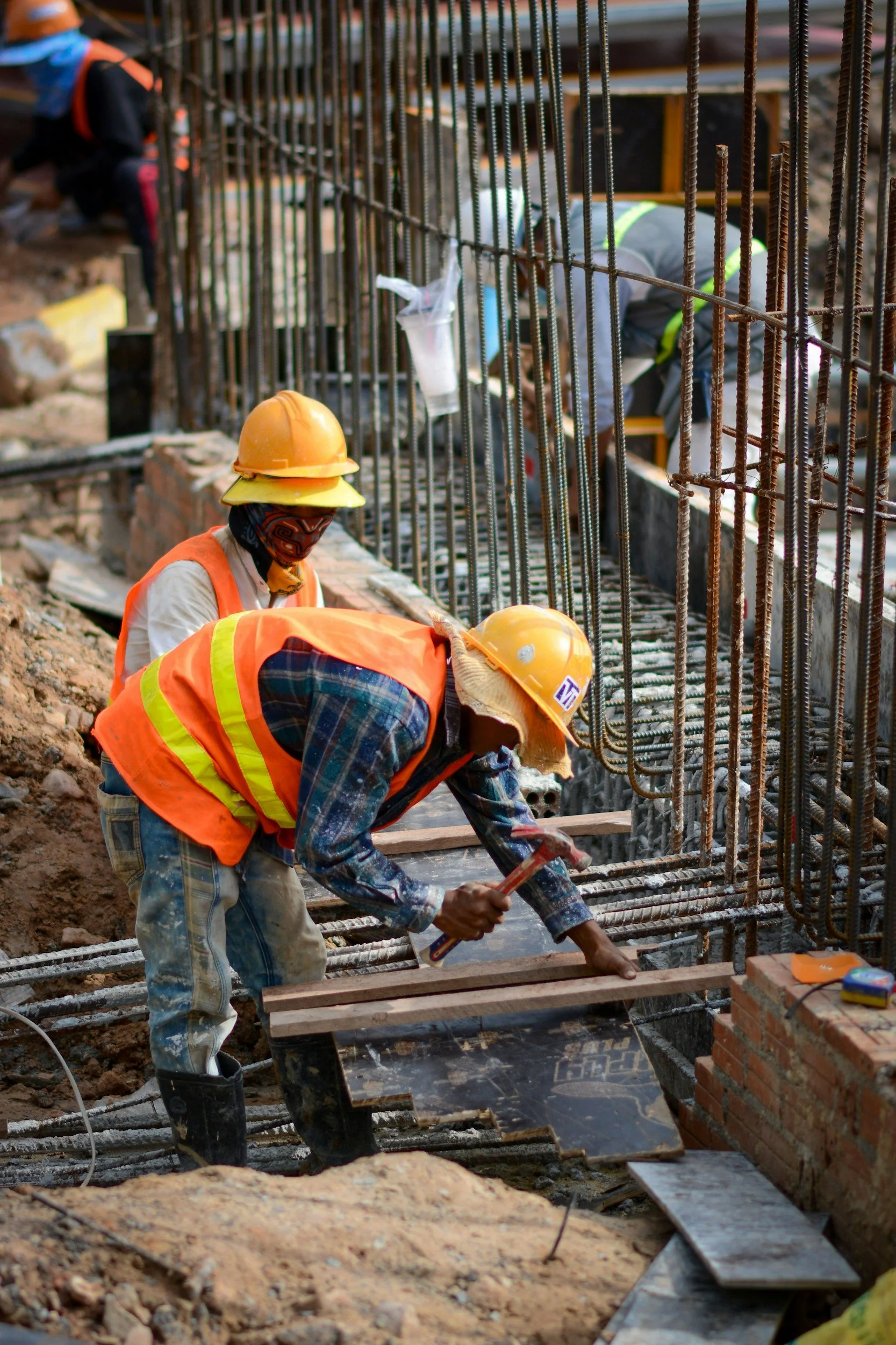 Construction workers wearing safety helmets and vests working on a building foundation, with steel rebar and brickwork visible.