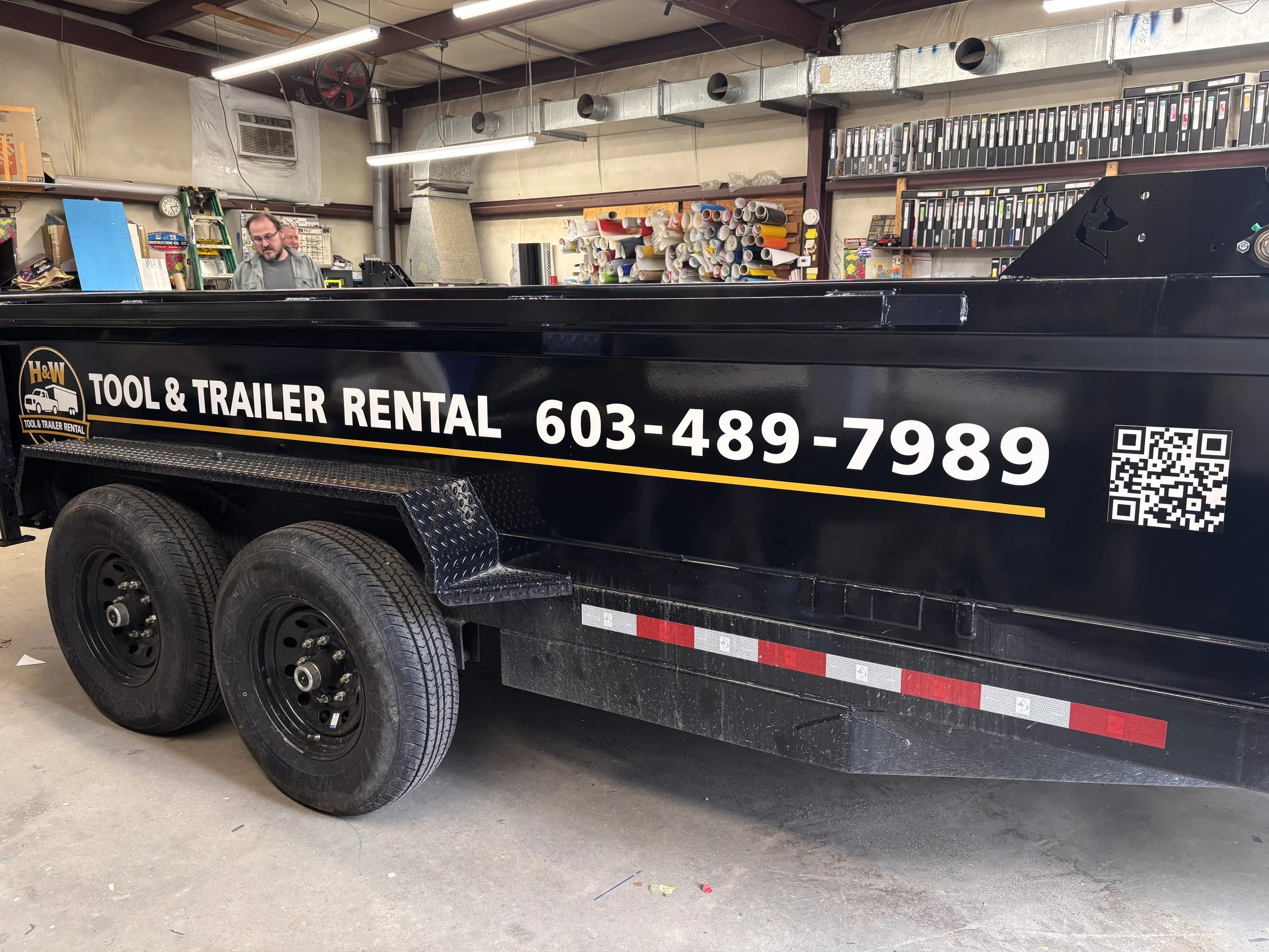 Black utility trailer with tool and trailer rental branding, contact number, QR code, and red and white reflective safety strip, parked indoors with shelves of rolled papers and binders in the background.