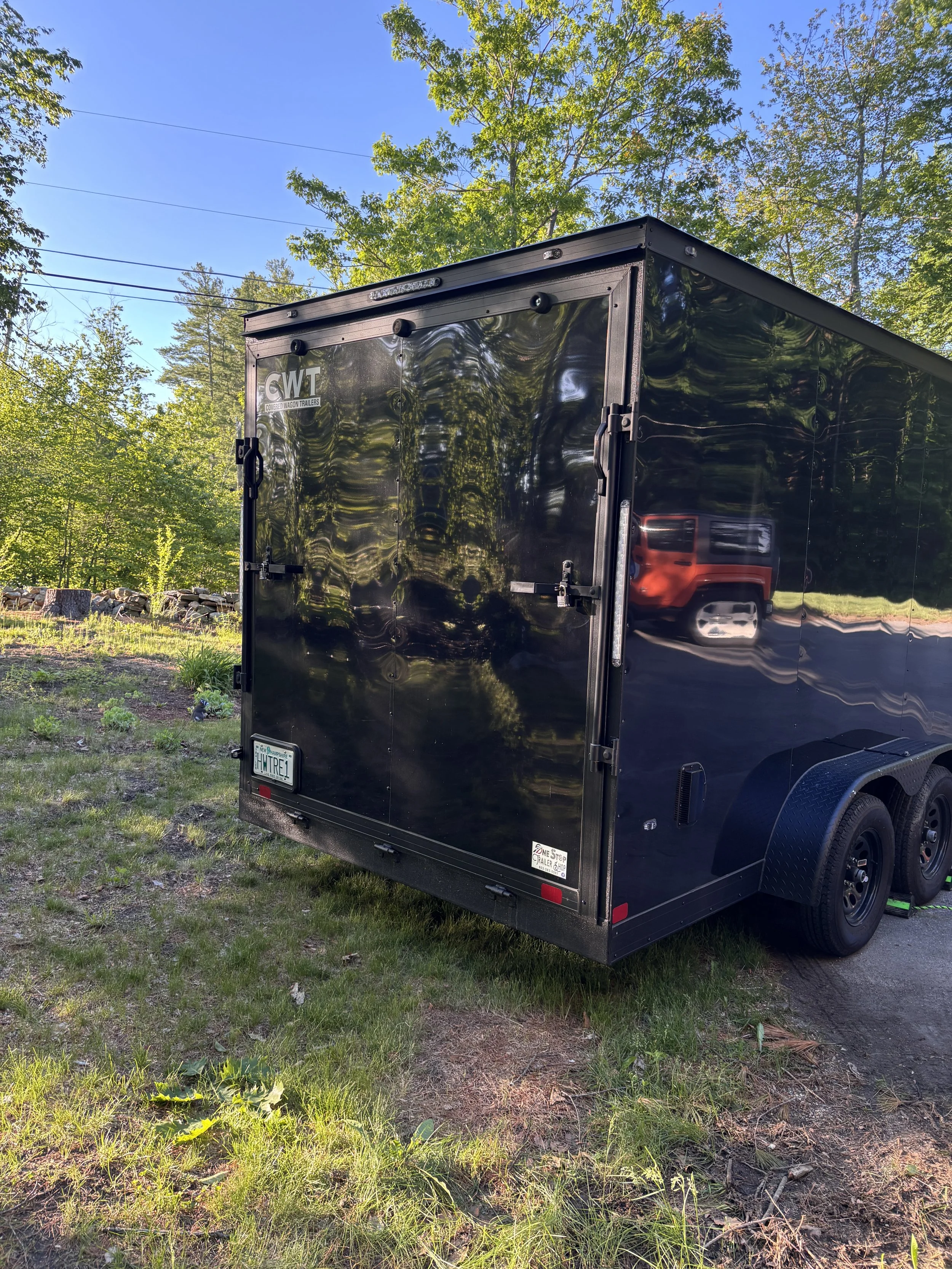A black enclosed trailer parked on grass with a reflection of trees and a vehicle on its shiny surface, under a clear blue sky.
