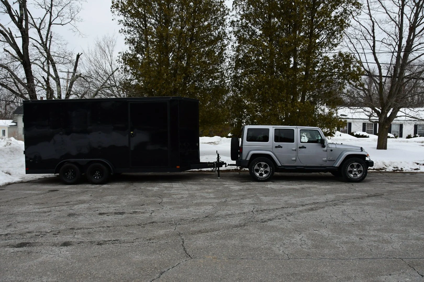 A silver Jeep SUV towing a large black enclosed trailer on a snowy street with leafless trees and houses in the background.