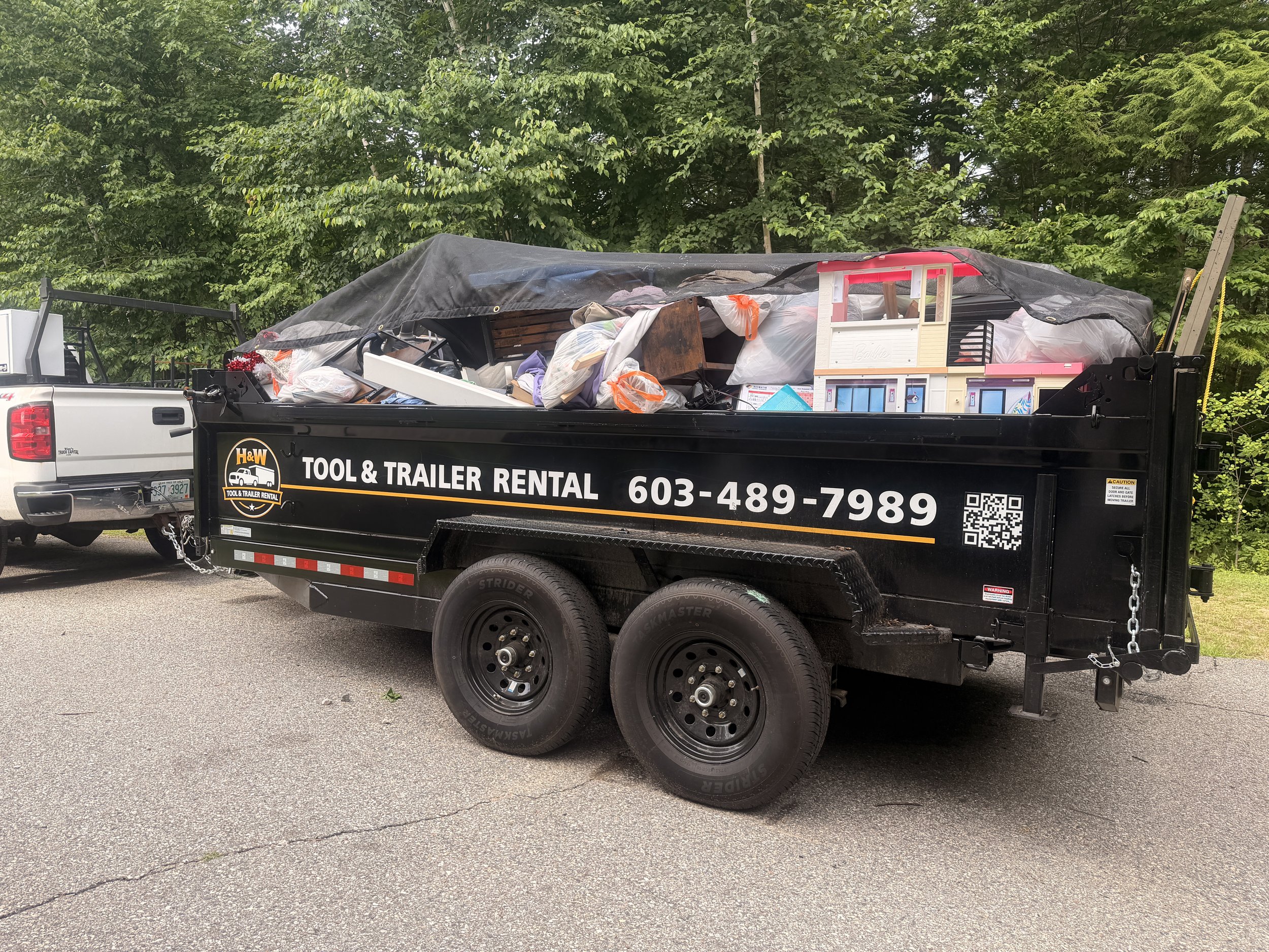 A black utility trailer attached to a white pickup truck, filled with various items, including a dollhouse, bags, and furniture, parked in front of green trees.