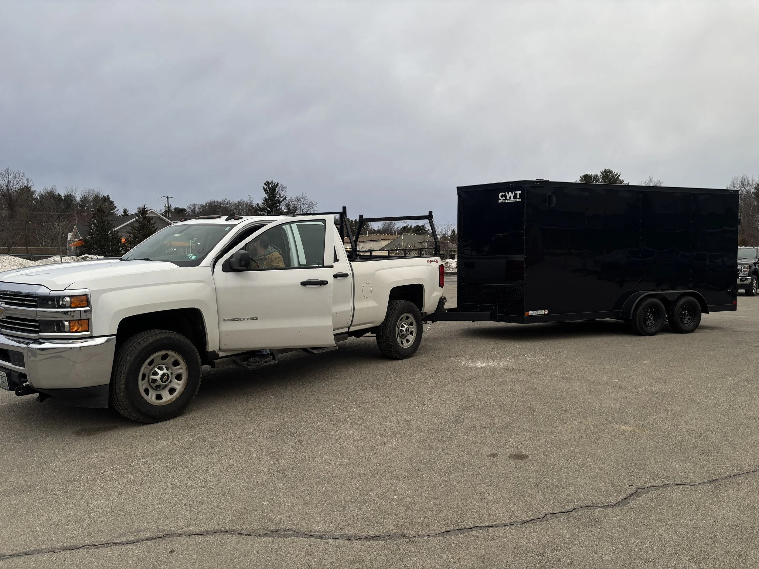 A white pickup truck with a black trailer attached. The trailer has the label 'CWT' on it. The truck is parked outdoors on a cloudy day with some snow on the ground. There are trees and houses in the background.
