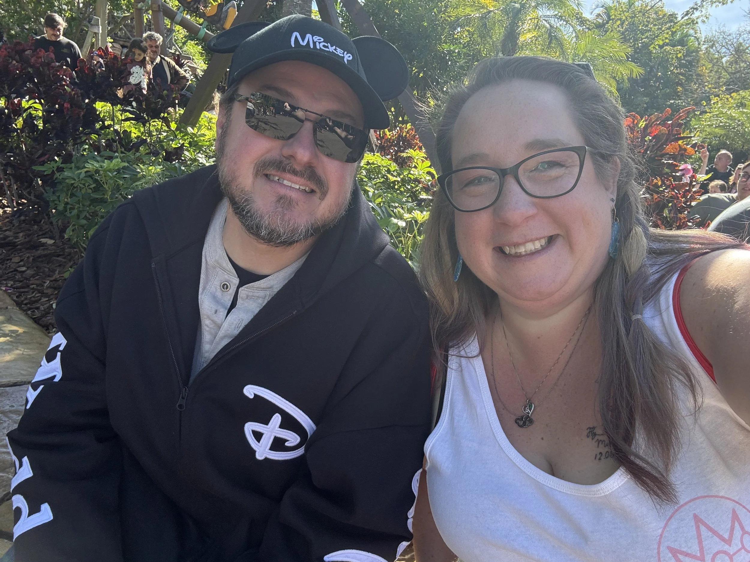 A smiling man wearing a black cap with Mickey Mouse ears and sunglasses, and a woman with glasses, smiling, in an outdoor setting with plants and trees in the background. There are other people in the distance.