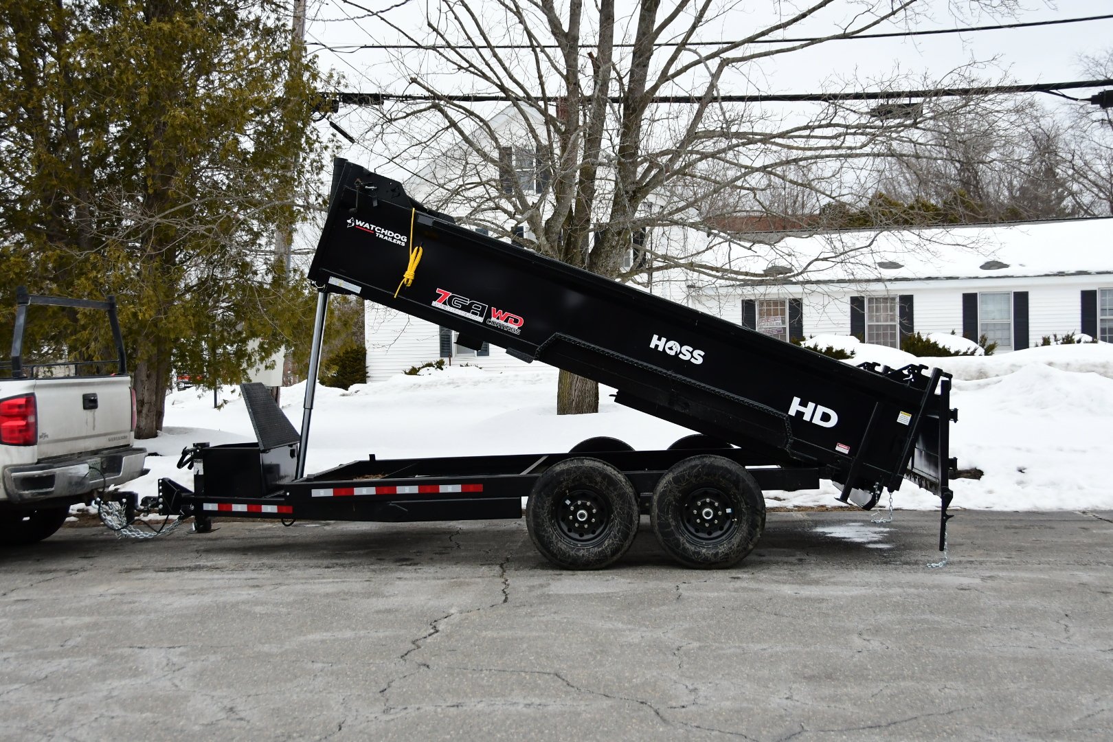 Black dump trailer attached to a pickup truck, parked on a snowy street with a background of trees and houses. Snow is piled on the ground and roofs.