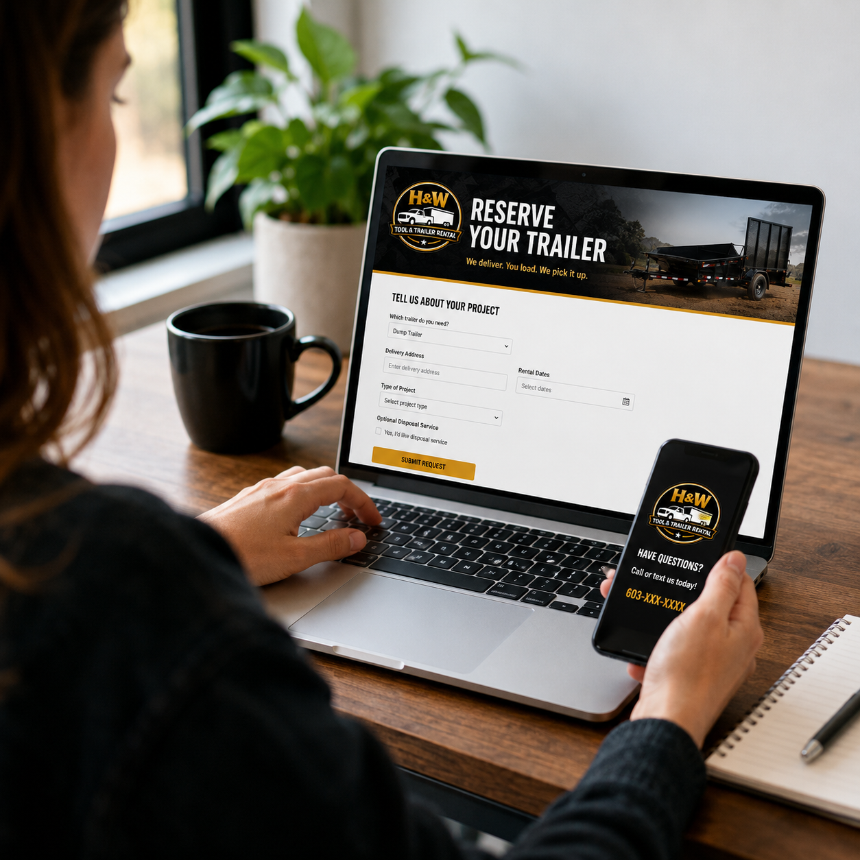 Person at a wooden desk holding a smartphone with a trailer rental business logo, looking at a laptop screen with a trailer reservation form, mug, potted plant, and notepad nearby.