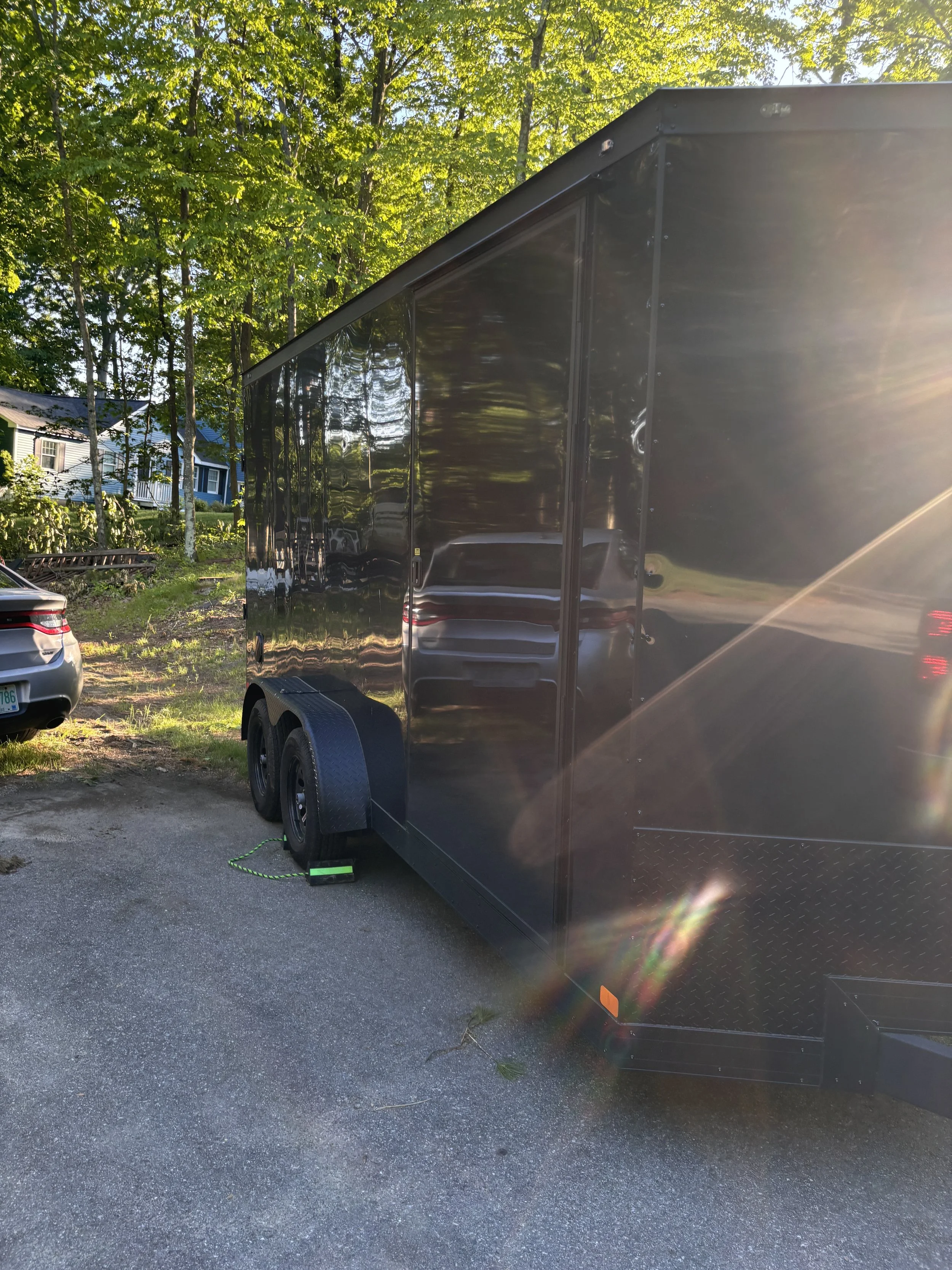 Photo of a black enclosed trailer parked on a driveway with a fence, grass, and houses in the background, and sunlight creating glare on the trailer.