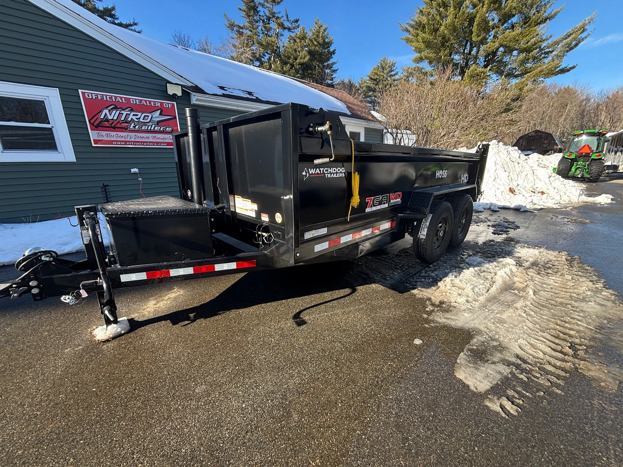 A black utility trailer is parked on a snow-covered driveway near a house with a sign that reads 'Official Dealer of Nitro Trailers.' A green tractor is visible in the background next to a large pile of snow. The scene is set on a bright, clear day with blue skies and trees in the background.