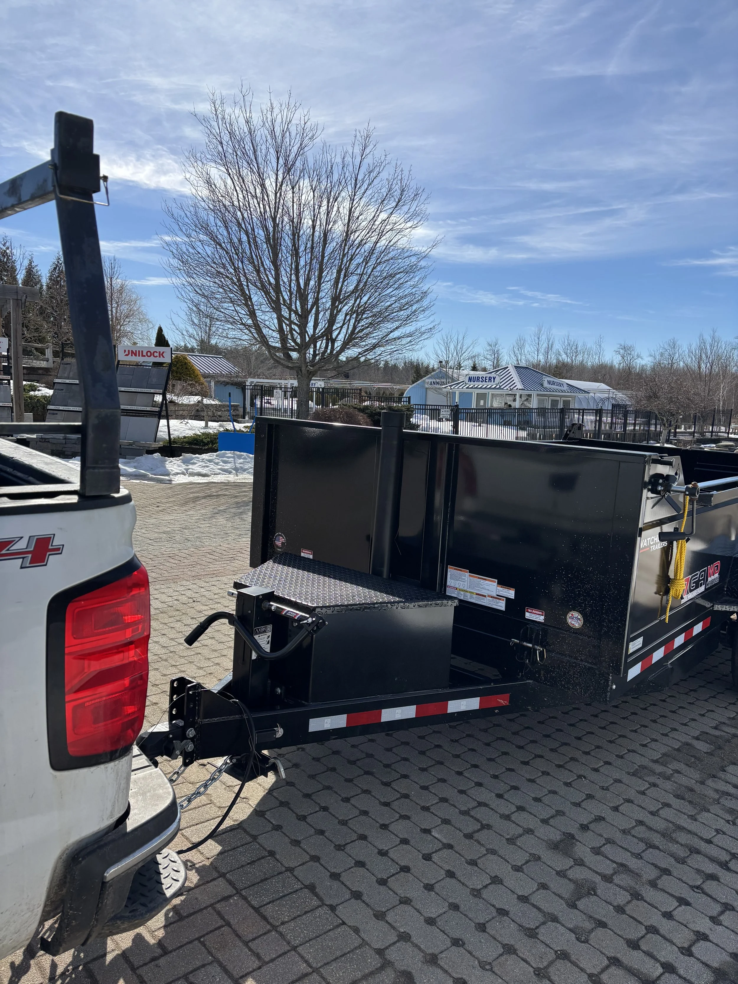 A trailer hitched to a pickup truck in a parking lot with snow on the ground, leafless trees, and building structures, under a partly cloudy blue sky.