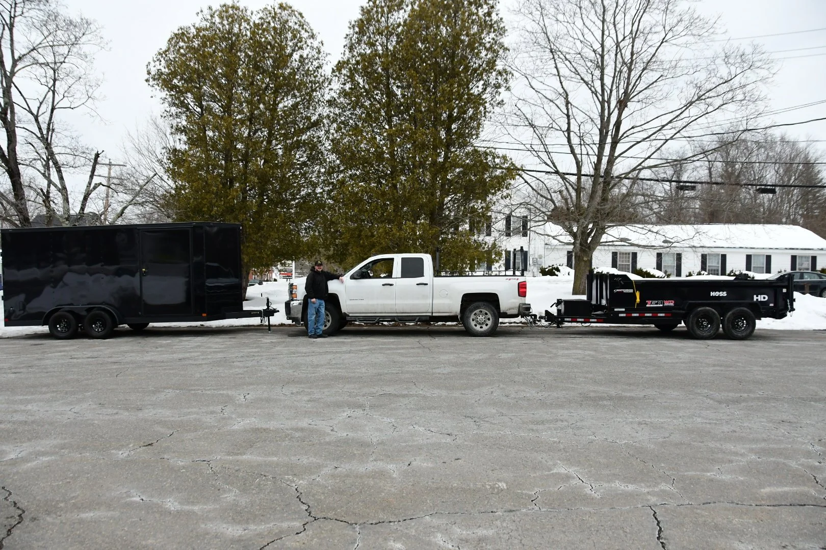 A man stands next to a white pickup truck attached to a black trailer with three axles, parked on a snowy street with leafless trees and a white house in the background.