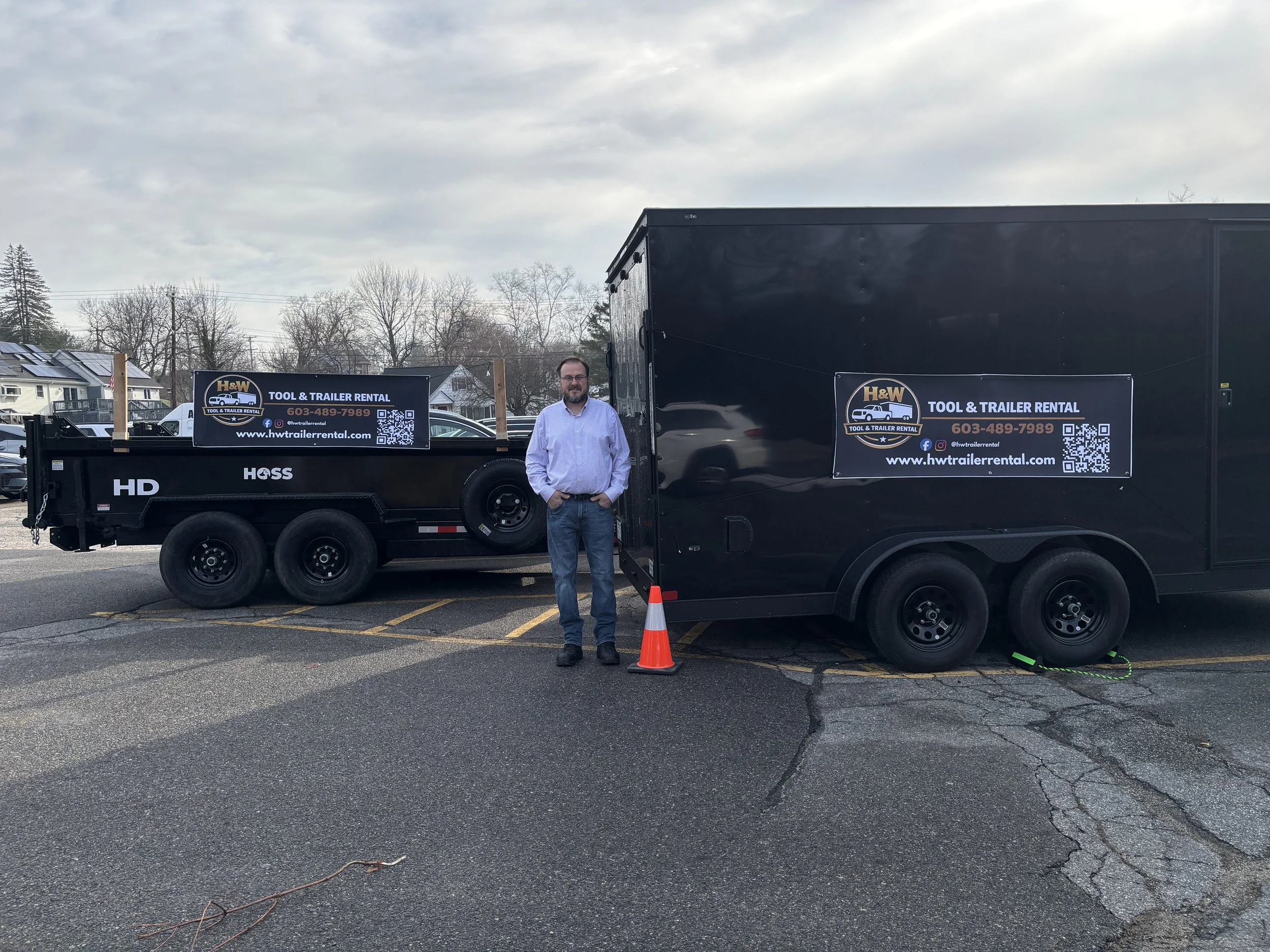 Man standing beside two black tool and trailer rental trailers parked on an asphalt lot, with trees and houses in the background and an overcast sky.