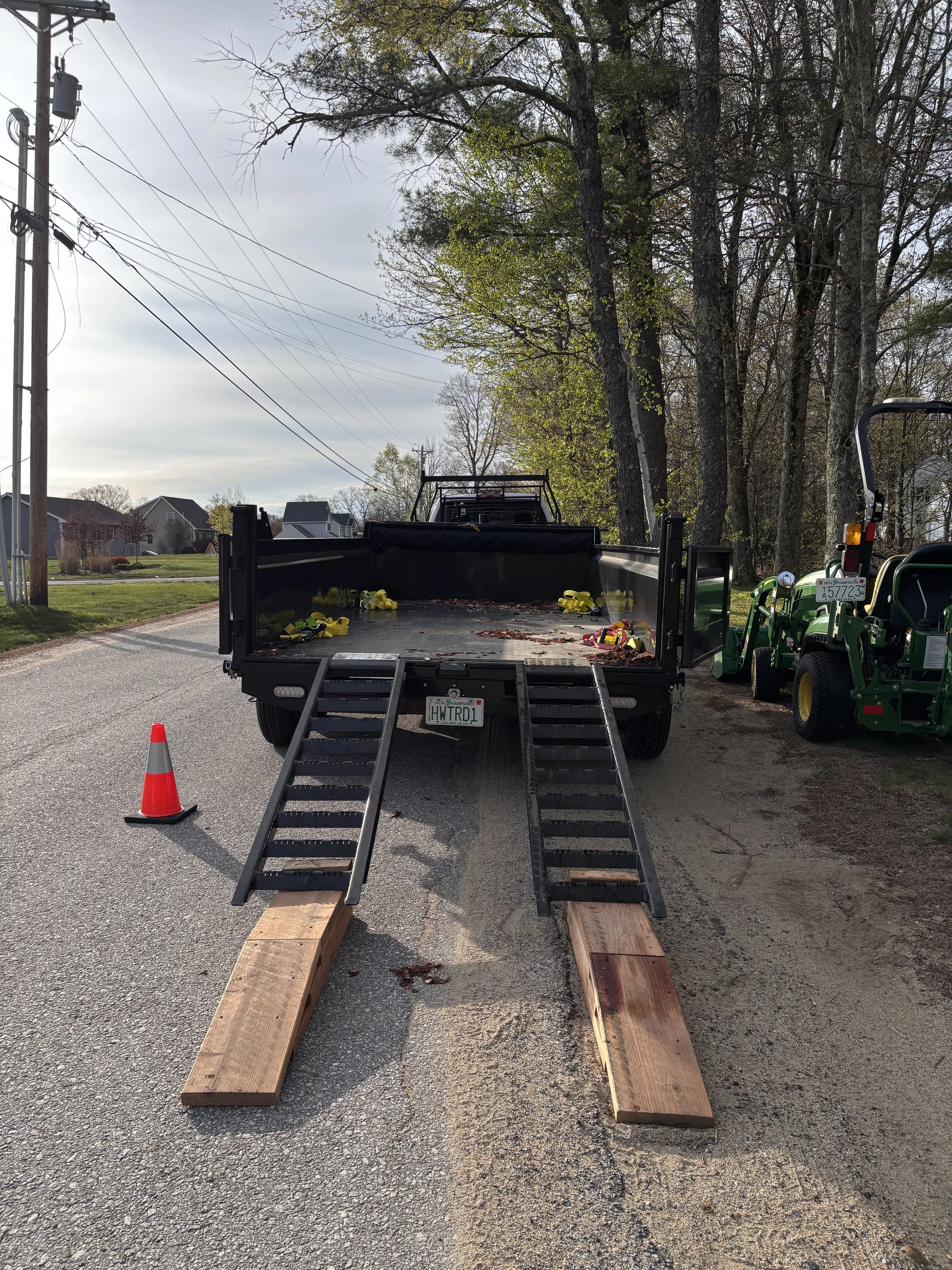 Open truck bed with wooden ramps leading to the ground, traffic cone to the left, and some yellow straps or harnesses inside the truck bed. A small green vehicle is parked to the right, and trees with green leaves are in the background.