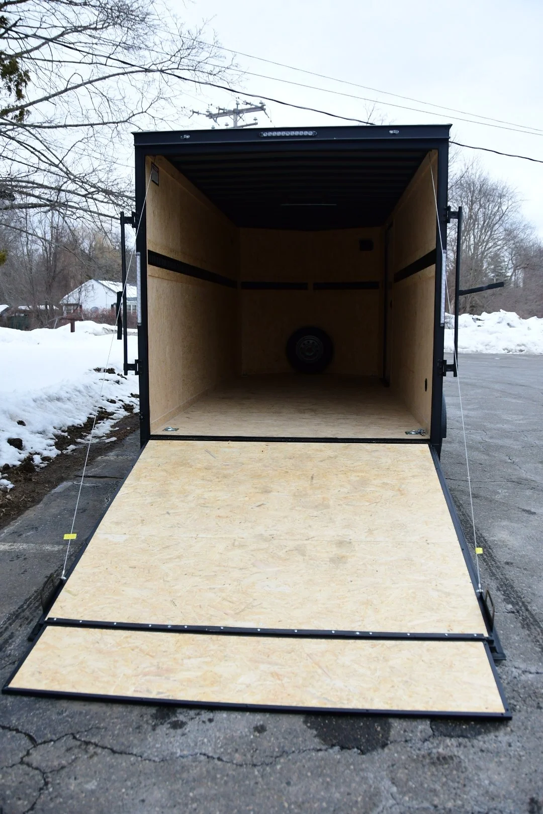 Open black enclosed cargo trailer with plywood interior and ramp door, parked on asphalt with snow on the ground and trees in the background.