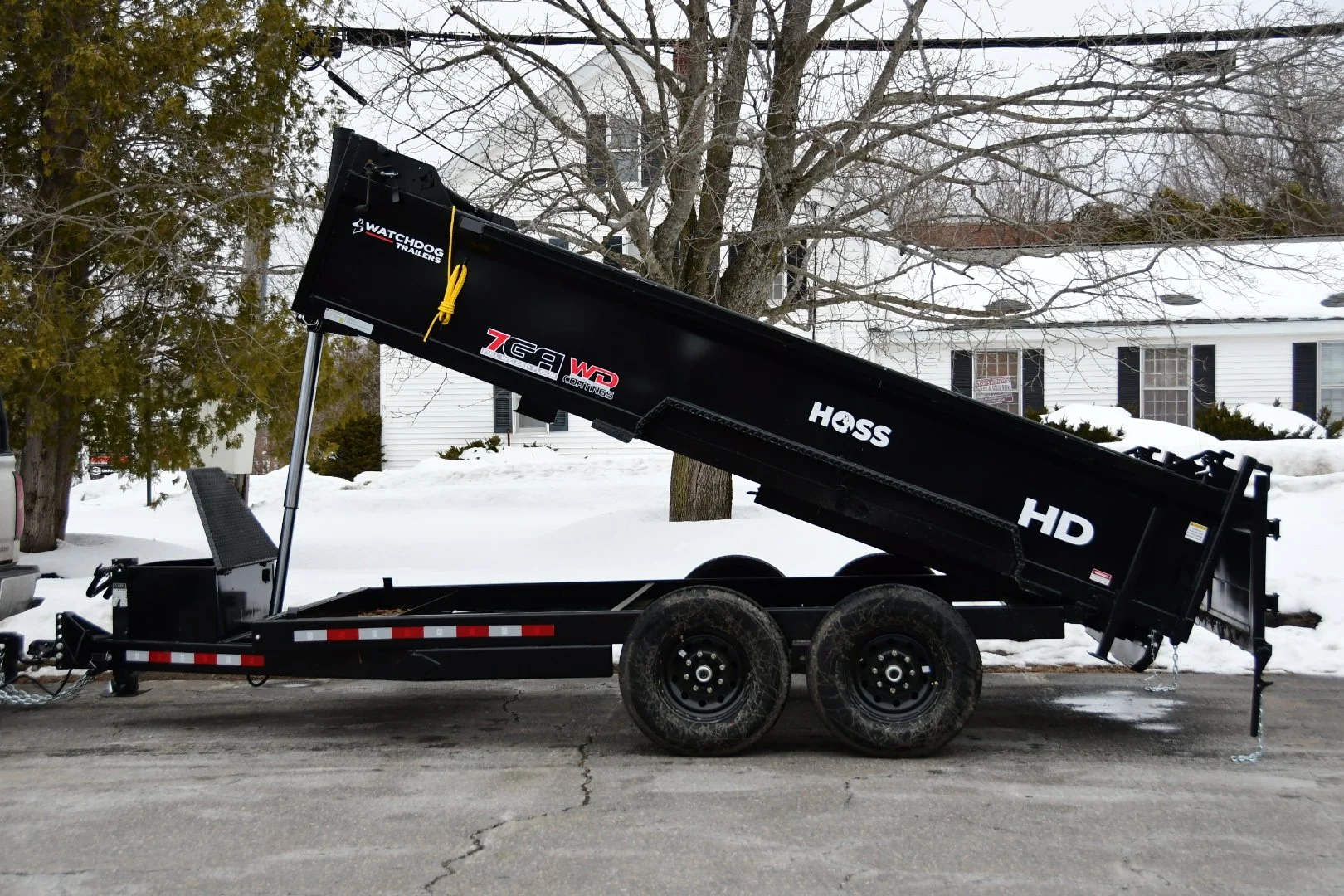 A black dump trailer with a hydraulic lift is parked on a snow-covered street, with houses and snow-covered trees in the background.